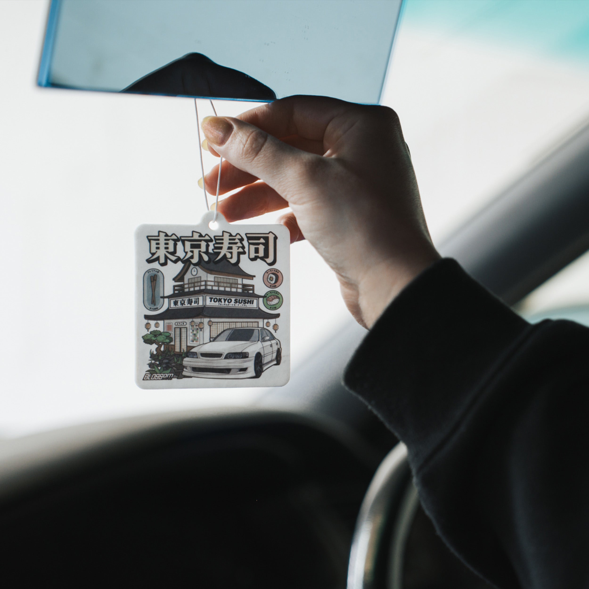 A person holds a Blossom Automotive 'Tokyo Sushi' Air Freshener - Green Tea, featuring a white car, a sushi restaurant, Japanese text, and "Tokyo Sushi", hanging inside a vehicle.