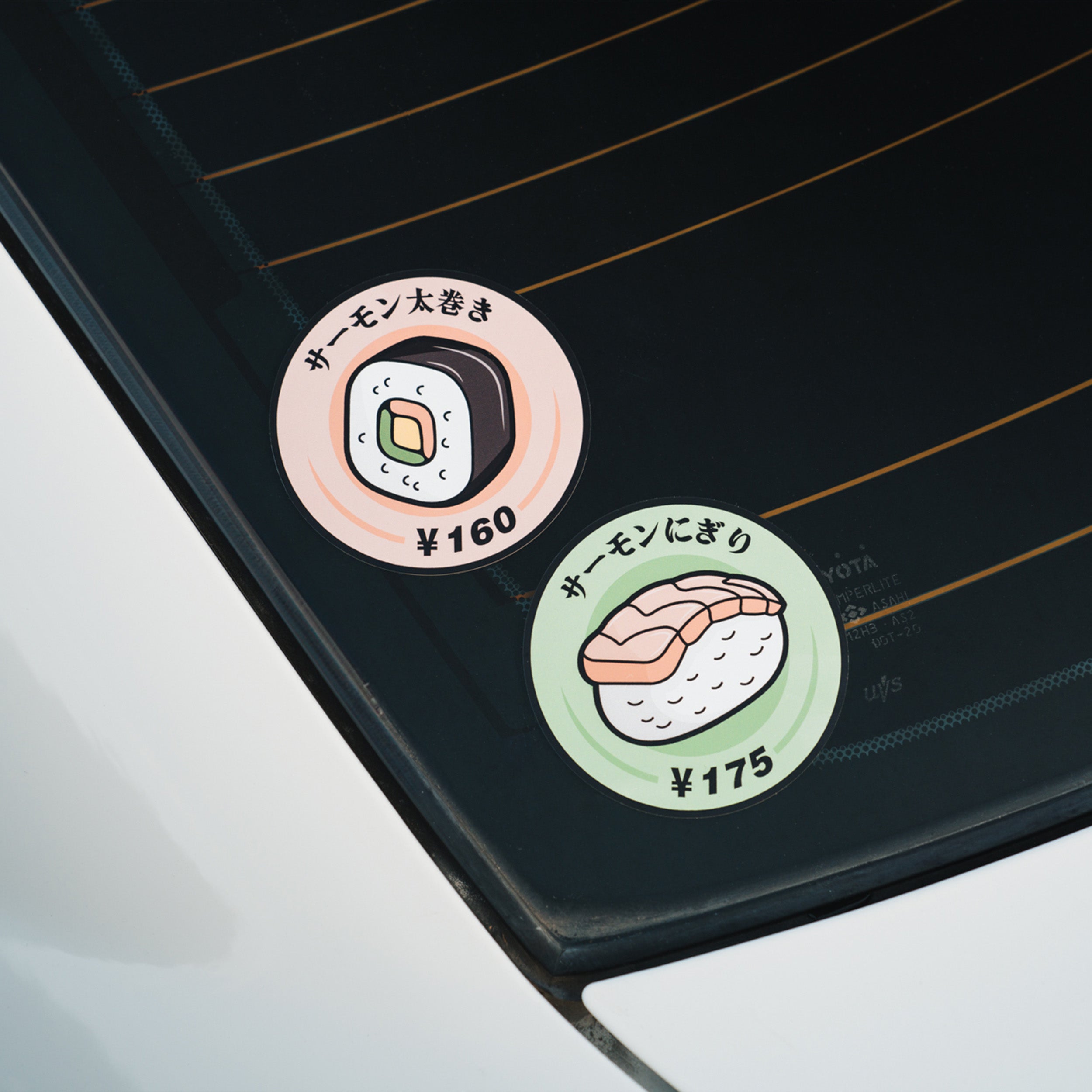 Two round Blossom Automotive stickers on a car window show illustrated sushi with Japanese text: one features the Nigiri Sushi - Slap Sticker priced at ¥175, and the other depicts a sushi roll sticker priced at ¥160.