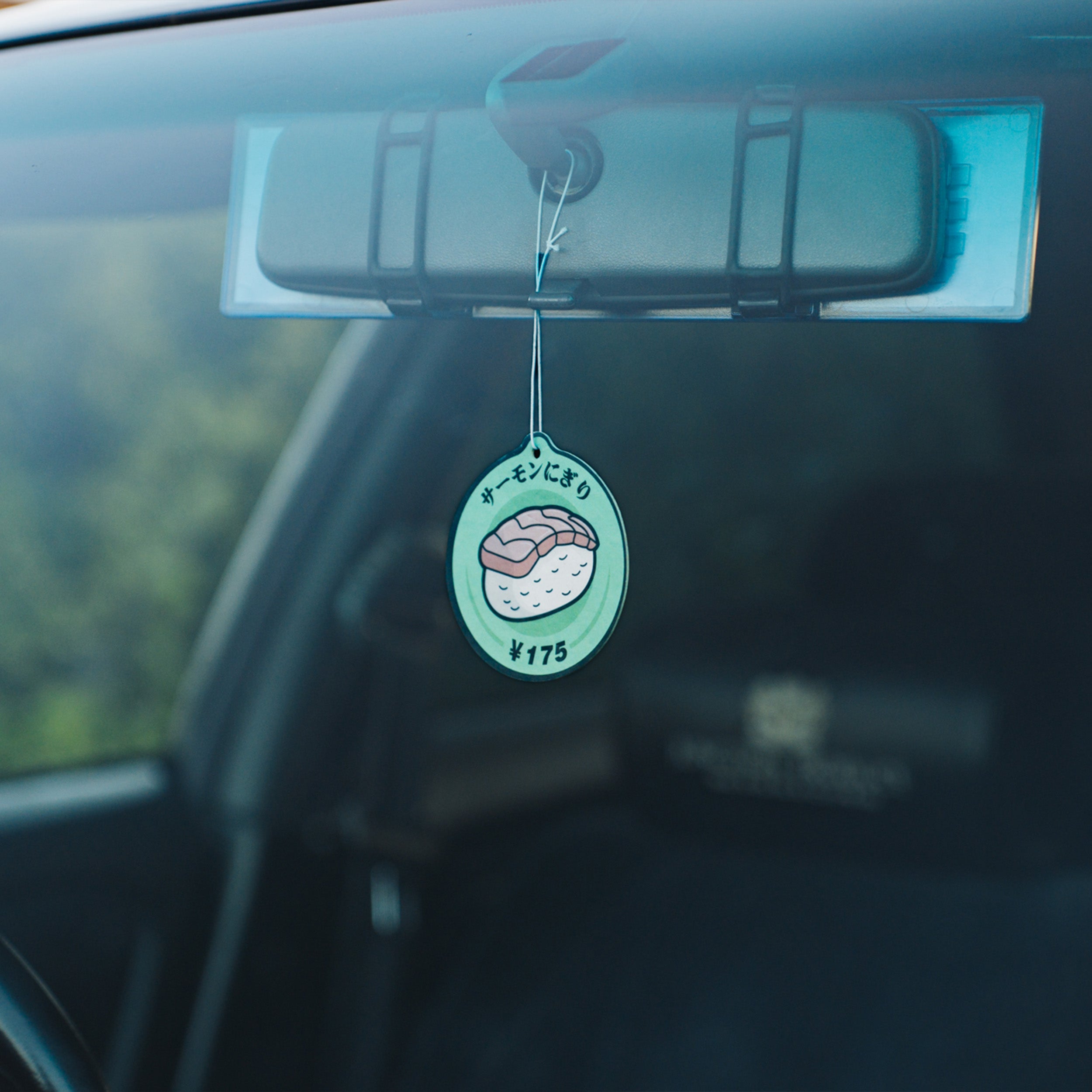 A 'Tokyo Sushi' Air Freshener - Mango by Blossom Automotive, priced at ¥175 and featuring Japanese text, hangs from a car’s rearview mirror as seen through the windshield.