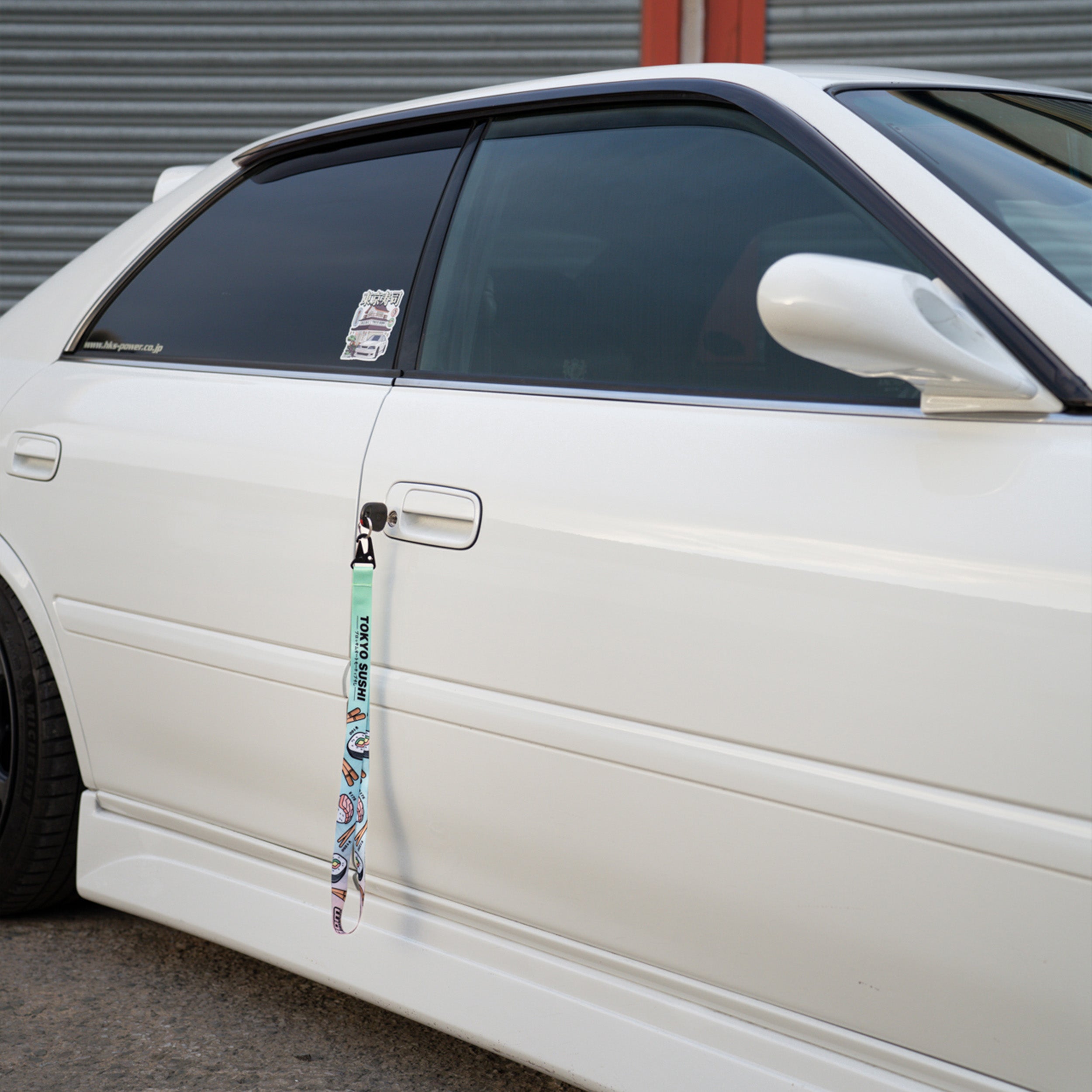 A close-up of a white car’s side door features the Blossom Automotive 'Tokyo Sushi Collection’ Lanyard hanging from the handle, with a sticker on the rear window. The car is parked beside a corrugated metal wall.