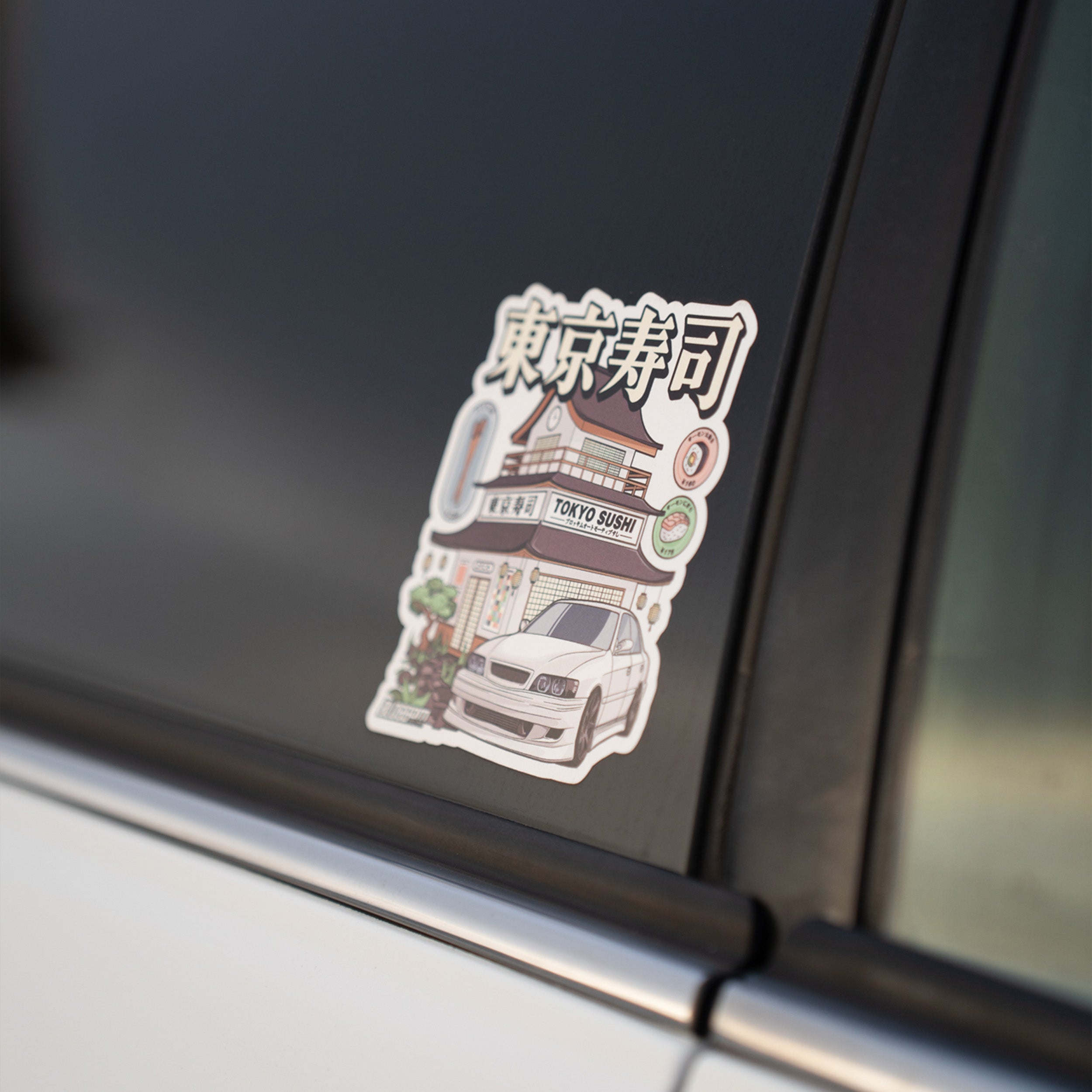 The Tokyo Sushi Collection - Slap Sticker by Blossom Automotive features a white car in front of a building labeled Tokyo Sushi, with Japanese characters above and decorative accents around the design.
