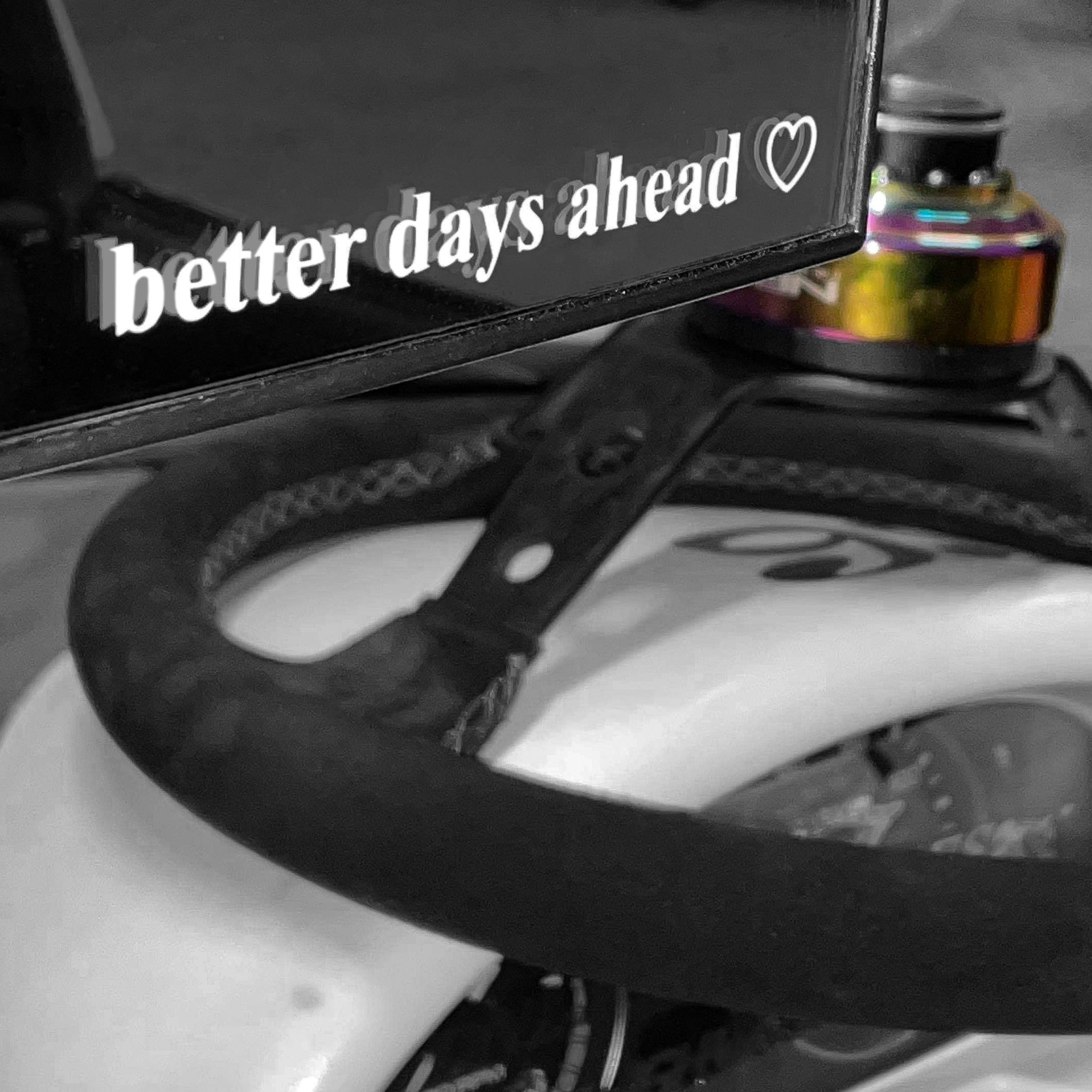 A close-up of a Blossom Automotive “Better Days Ahead” die-cut sticker shows the phrase and a heart in white on a black bicycle mirror, with the mostly black-and-white image highlighting one colored bike part.