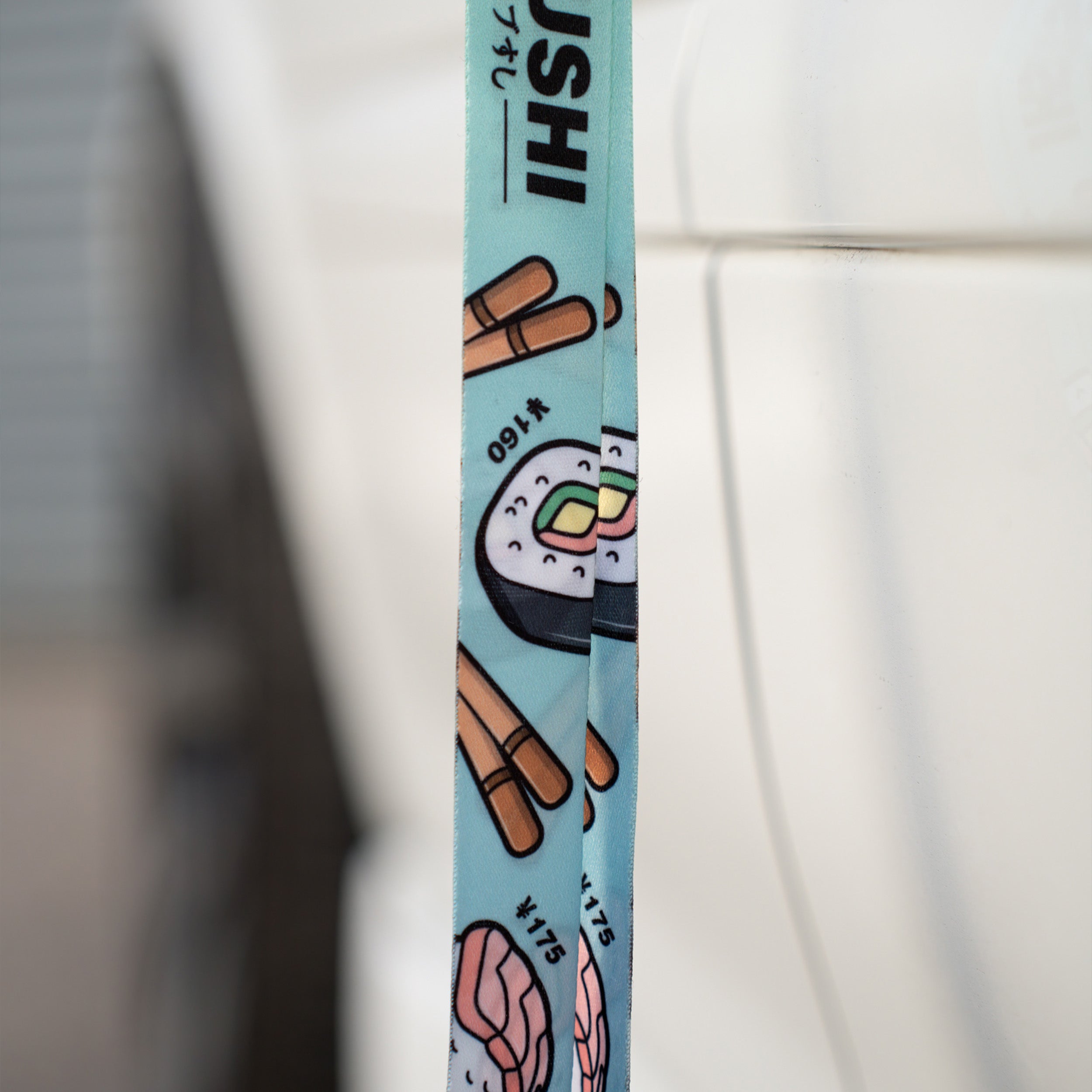 A close-up of Blossom Automotive’s ‘Tokyo Sushi Collection’ Lanyard, featuring blue fabric with cartoon sushi rolls, chopsticks, nigiri, and the word SUSHI, hanging in front of a white surface.