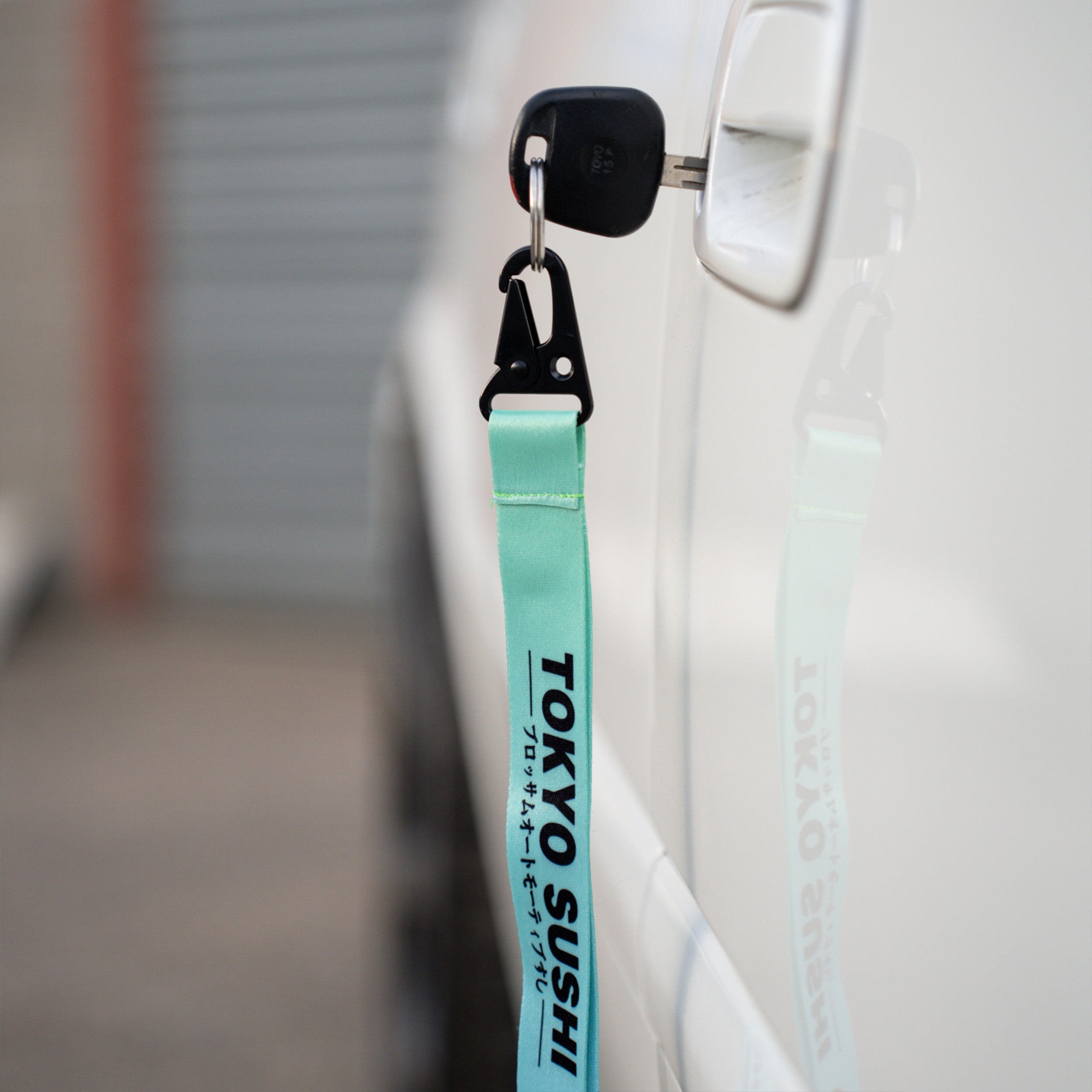 A car key with a turquoise Blossom Automotive ‘Tokyo Sushi Collection’ lanyard, featuring English and Japanese text, is inserted into the door of a white vehicle, its reflection visible on the car’s surface.