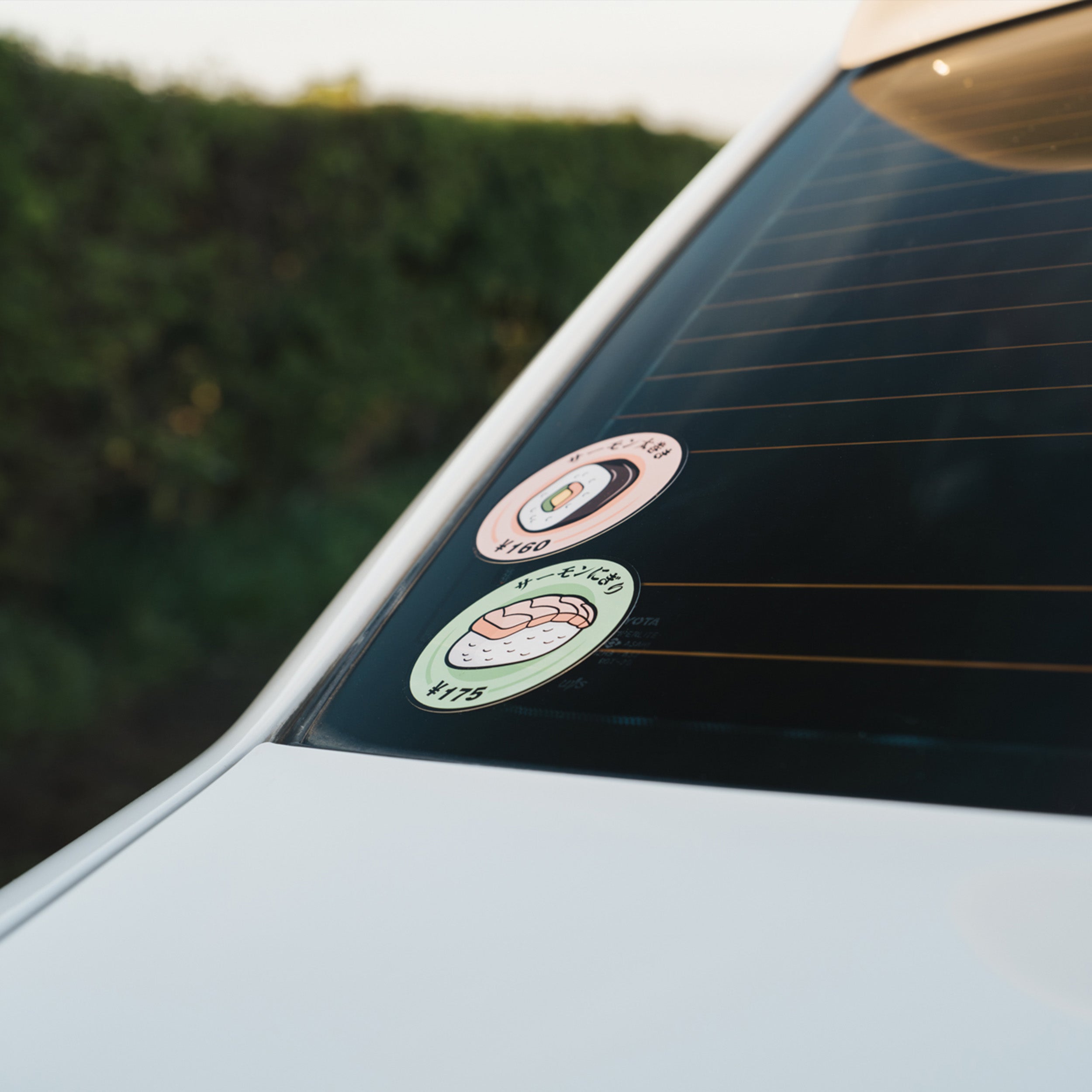 A Blossom Automotive Nigiri Sushi - Slap Sticker and a donut sticker decorate the rear window of a white car, set against lush greenery.