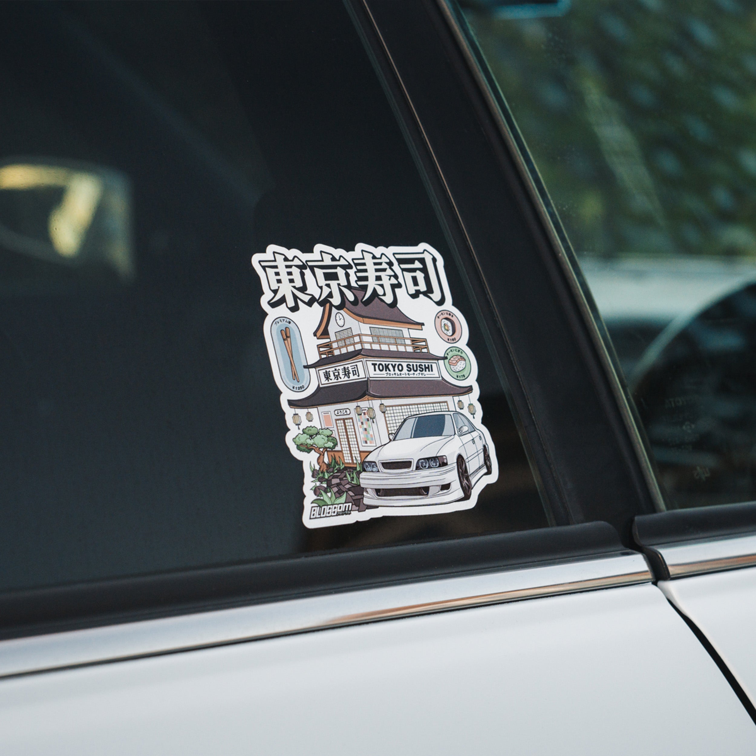 The Blossom Automotive Tokyo Sushi Collection - Slap Sticker shows a Japanese sushi restaurant with a white car parked in front. Japanese characters and “Tokyo Sushi” appear on the sign above the building.