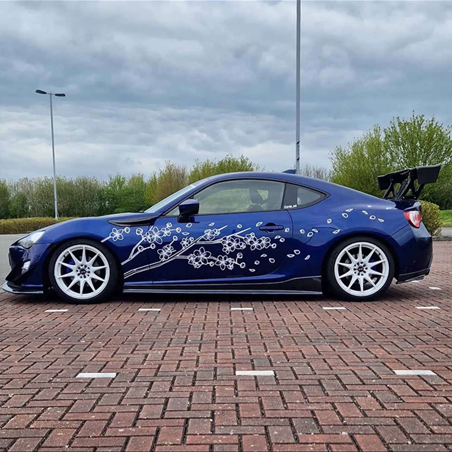 Blue sports car with white floral design on a brick road under a cloudy sky.