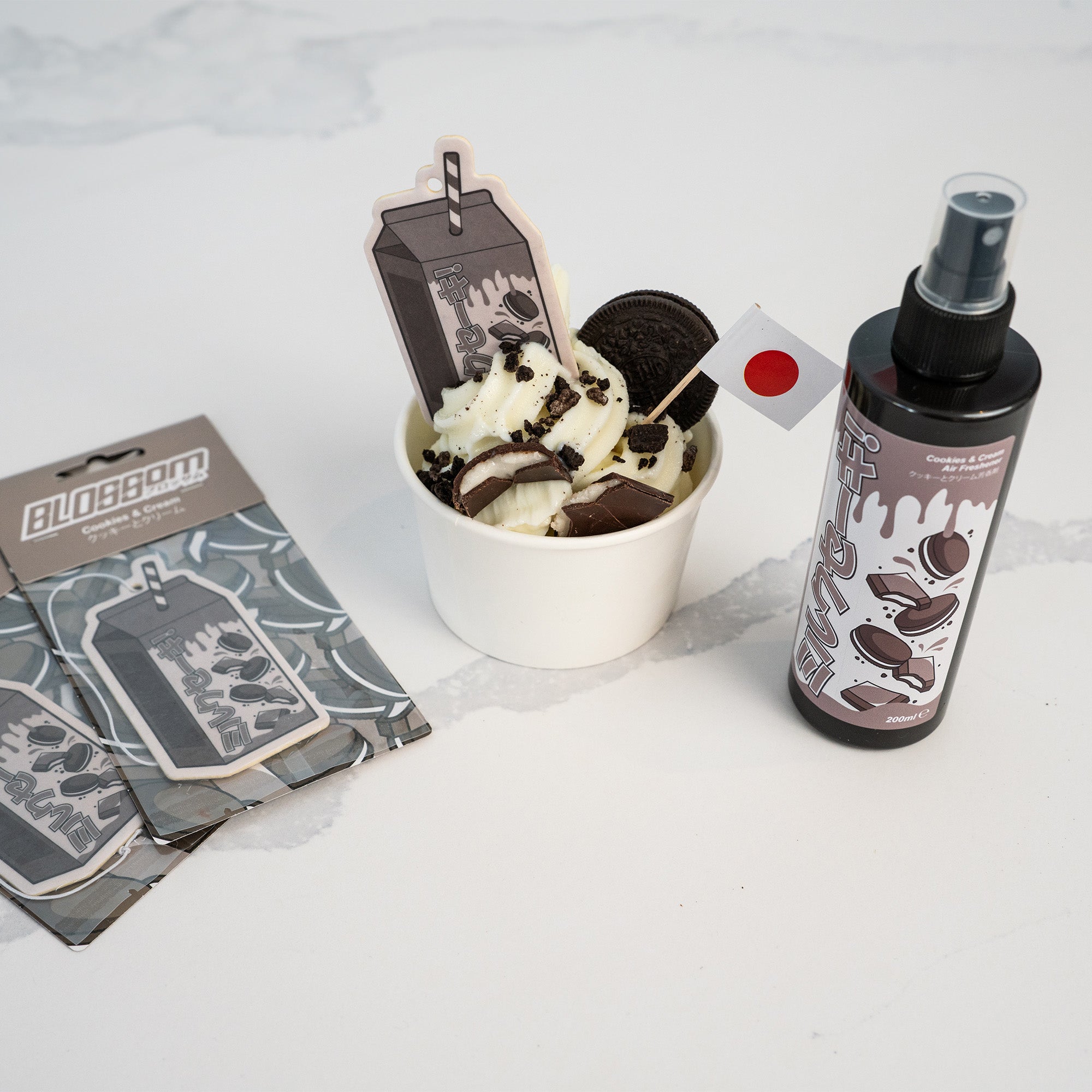 A 'The Milkshake Collection' Cookies & Cream Car Spray Air Freshener by Blossom Automotive is displayed beside card packs with matching designs and a soft-serve ice cream cup topped with chocolate, an Oreo cookie, and a small Japanese flag.