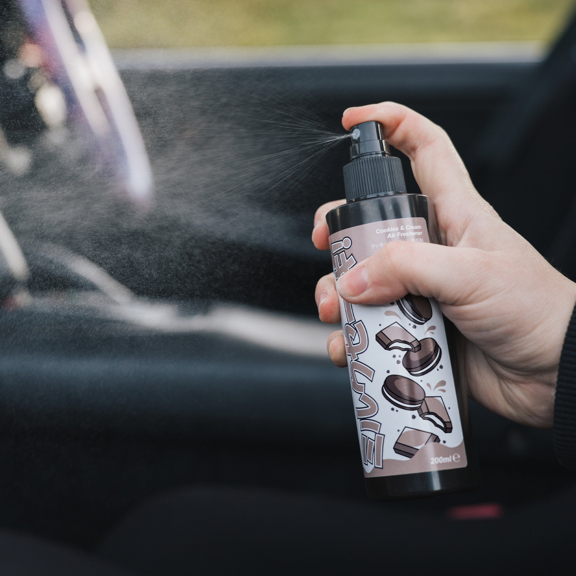 A hand sprays Blossom Automotive’s 'The Milkshake Collection' Car Spray Air Freshener - Cookies & Cream 200ml inside a car, releasing a fine mist, while the blurred steering wheel and seat appear in the background.