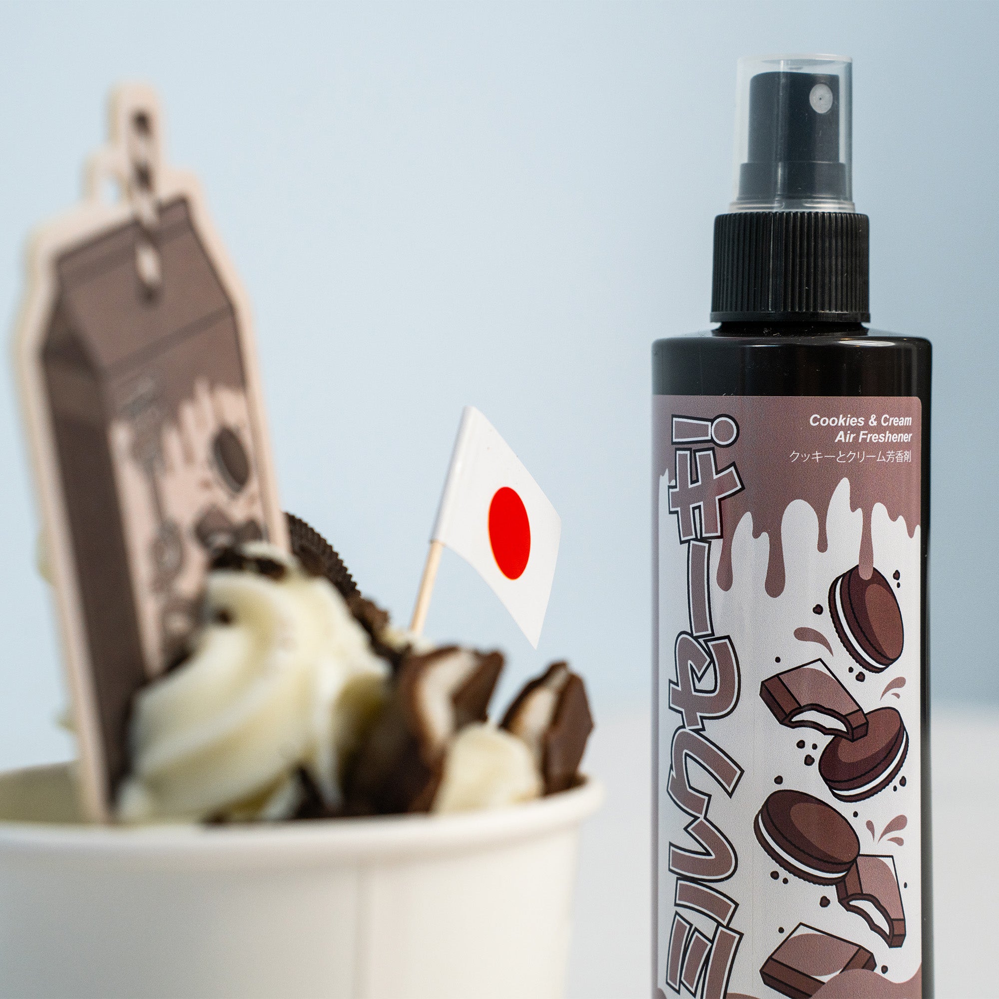 A cup of cookies and cream dessert with a Japanese flag sits beside a Blossom Automotive 'The Milkshake Collection' Car Spray Air Freshener Bundle (2x200ml) featuring cookies on the label, both displayed against a light background.