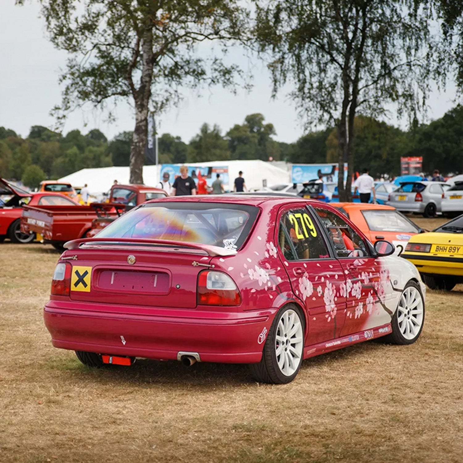 Red car with cherry blossom livery at a car show with trees and other cars in the background