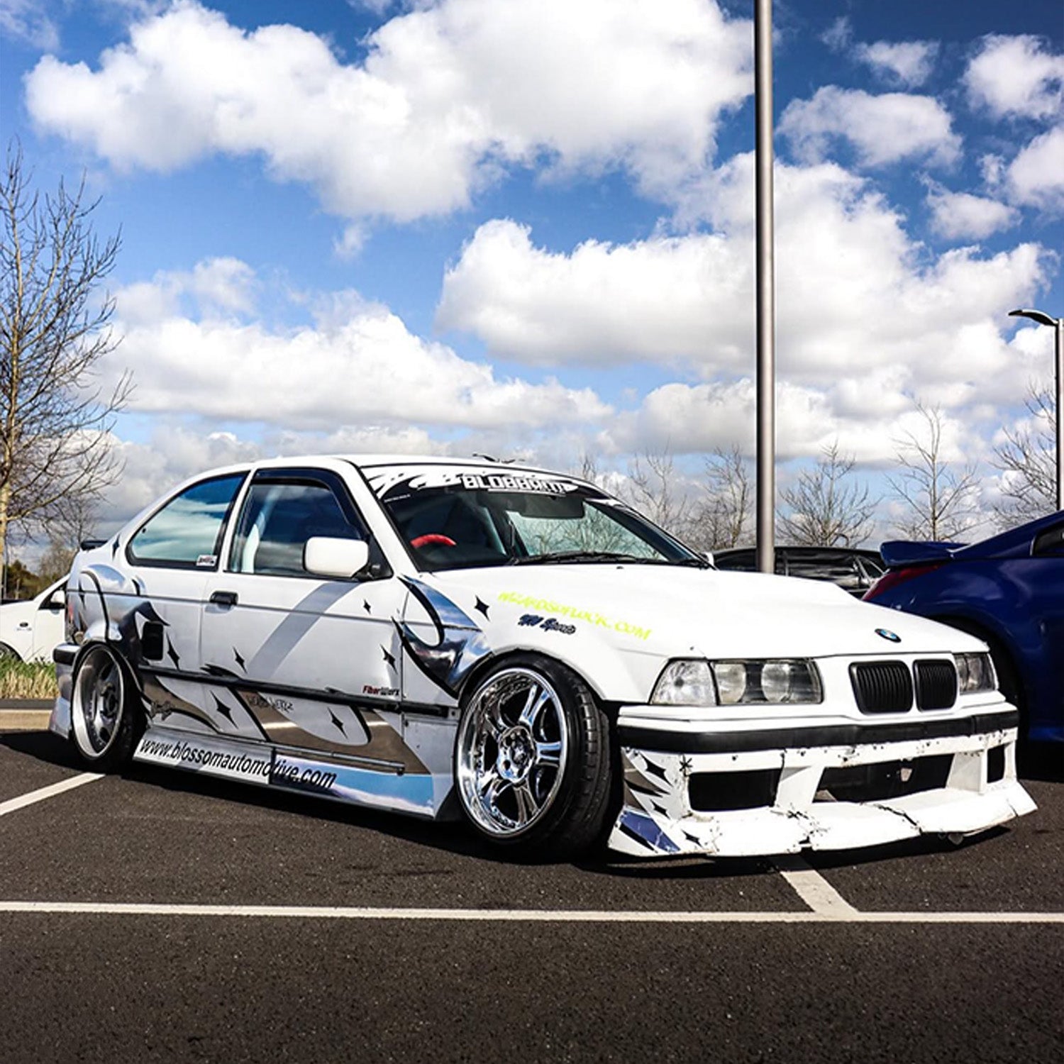 White BMW car with Zoku tribal livery on a parking lot with a blue sky.