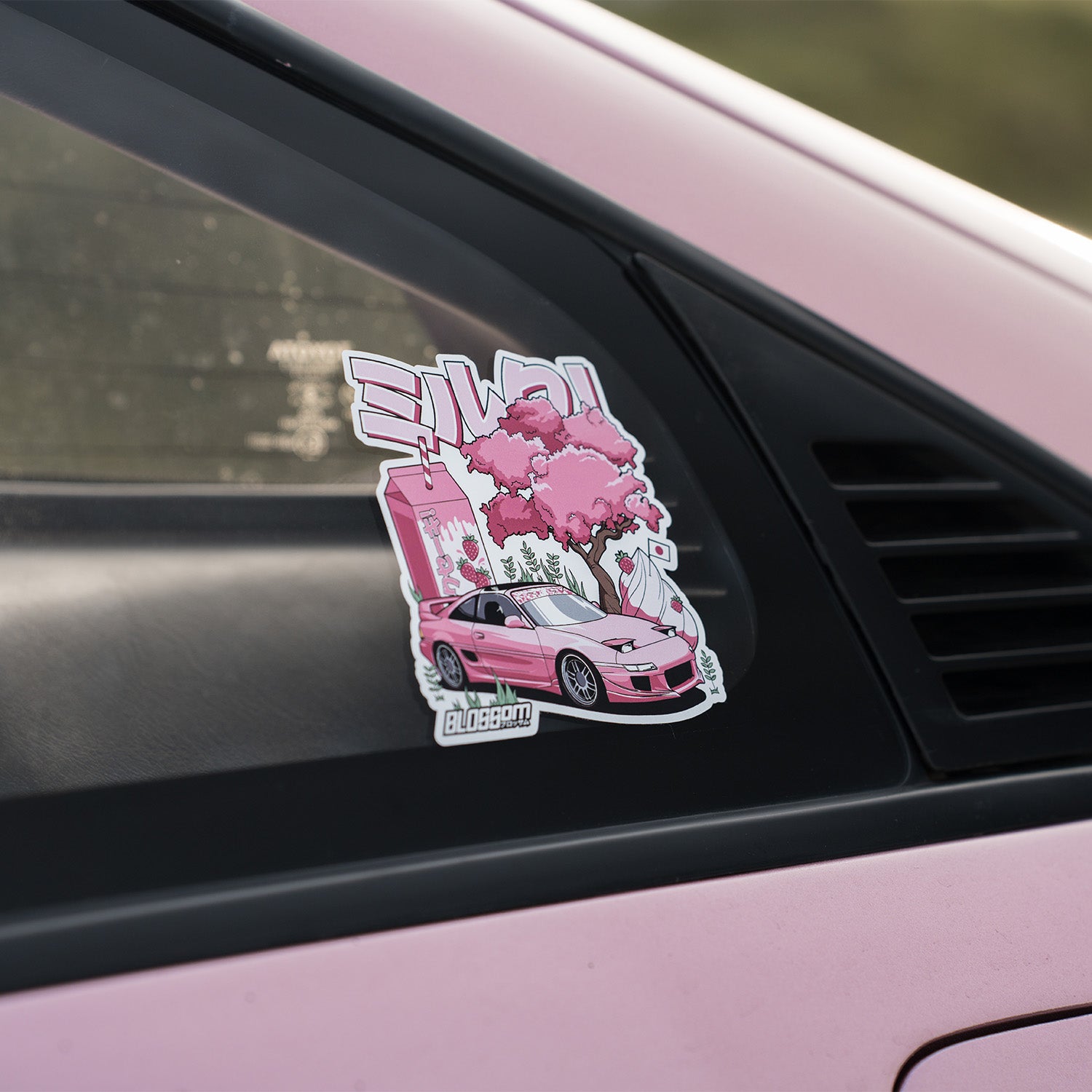 The Milkshake Collection - Slap Sticker by Blossom Automotive features a pink sports car, cherry blossom tree, and Japanese text, adding a playful anime vibe to your car’s rear window.
