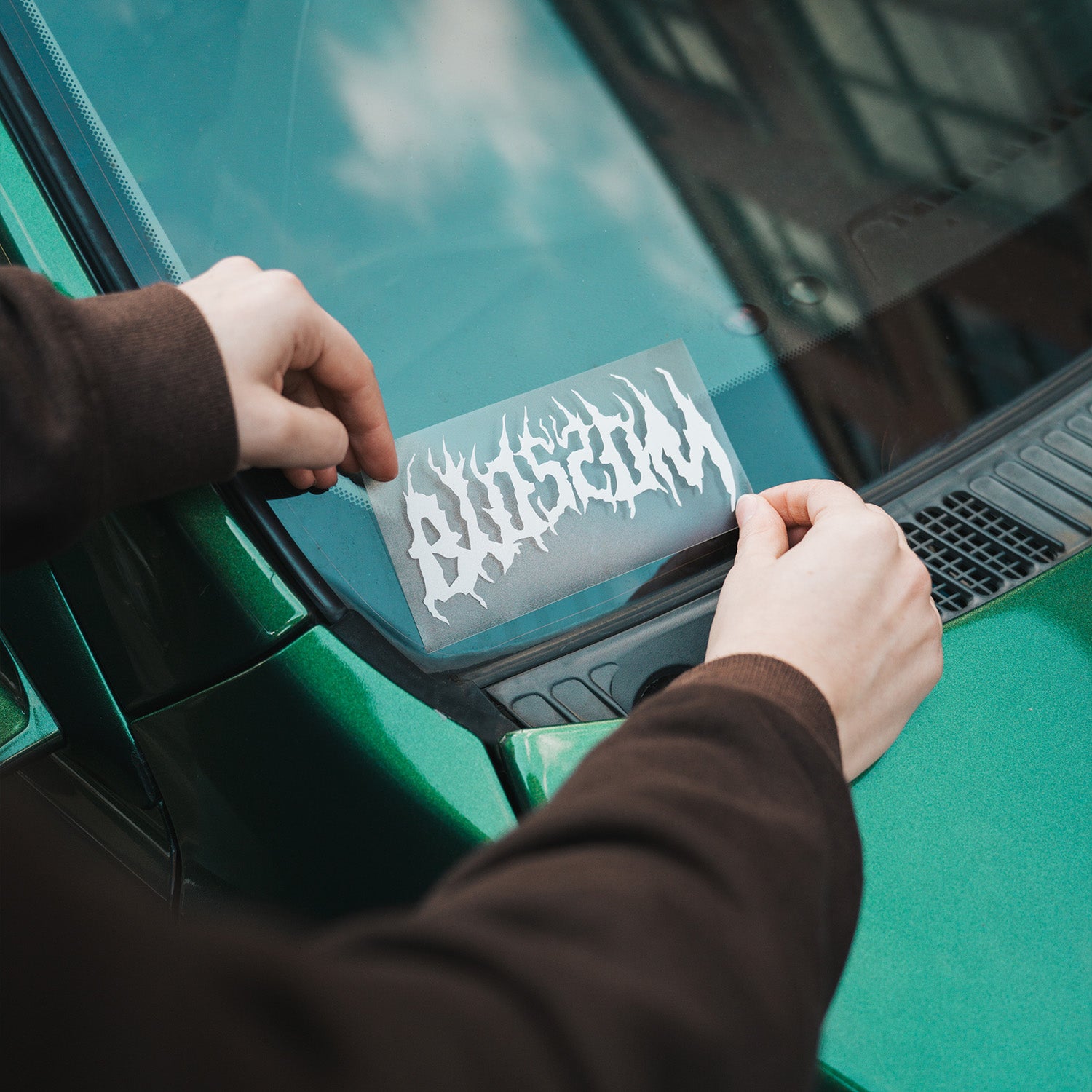 Someone applies a Blossom Automotive Blossom Skate Logo die cut sticker, featuring a stylized, flame-like design, to the windshield of a green car.