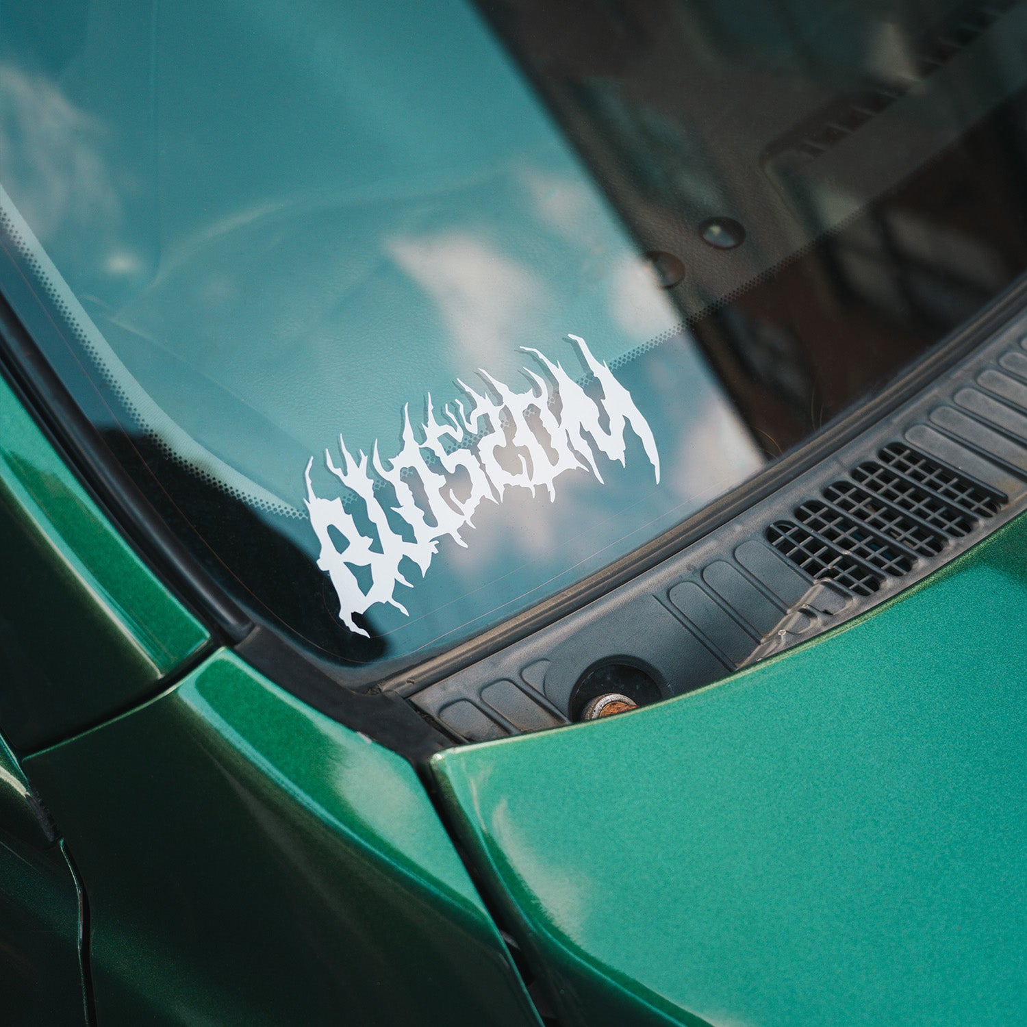 A Blossom Automotive Blossom Skate Logo - Die Cut Sticker in white with a jagged, flame-like font is displayed on the windshield of a green car.