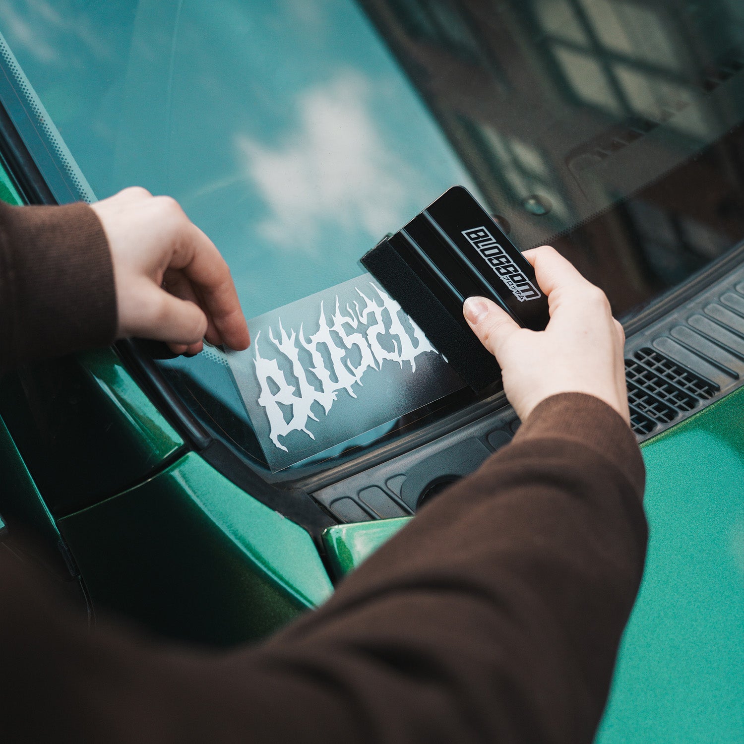 A person applies the Blossom Skate Logo - Die Cut Sticker by Blossom Automotive, featuring jagged, flame-like white lettering, to the windshield of a green car using a black plastic tool.