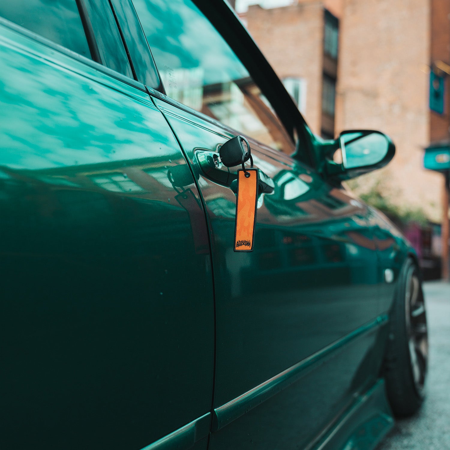 A close-up of a teal car parked on the street, with a black key and an orange Blossom Automotive 'The Skate Collection’ Flight Tag hanging from the driver’s side door handle. The window reflects blue sky and clouds.