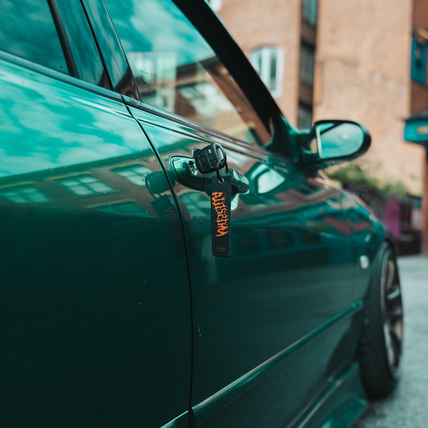 A ‘The Skate Collection’ Flight Tag by Blossom Automotive hangs from a car key inserted into the door lock of a shiny green car parked on a city street, with blurred buildings in the background.