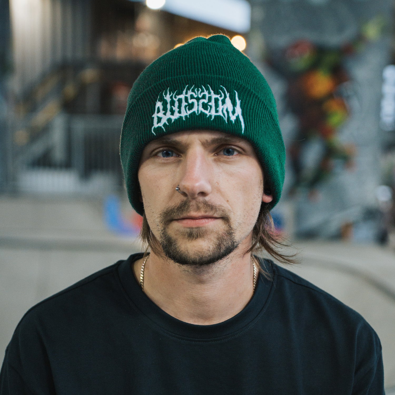 A man with a mustache and goatee, wearing Blossom Automotive's 'The Skate Collection’ Beanie in bottle green and a black shirt, looks directly at the camera inside an indoor skate park.