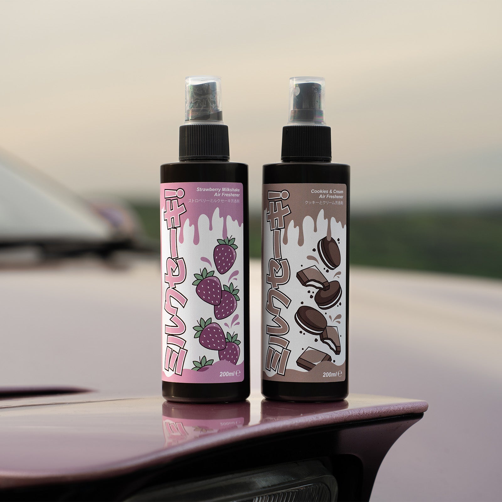 Two playful spray bottles from Blossom Automotive’s Milkshake Collection sit on a pink car: one labeled Strawberry Milkshake with strawberries, the other featuring cookies and cream illustrations as Cookies & Cream Car Spray Air Freshener (200ml).
