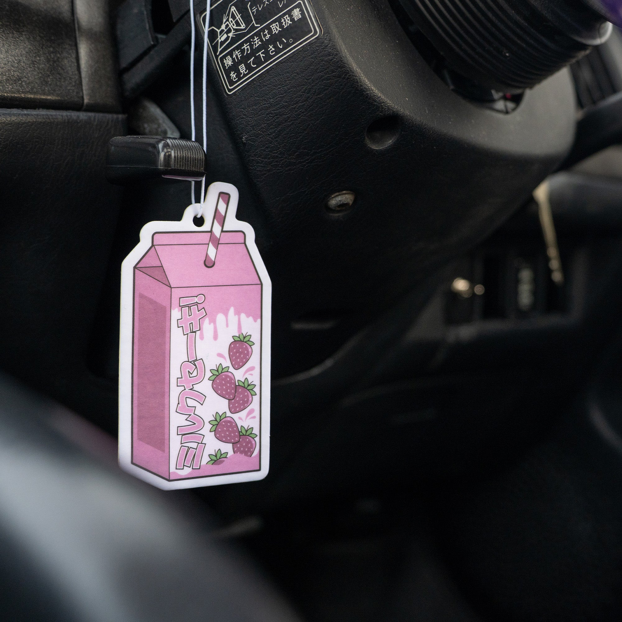 A ‘The Milkshake Collection’ Strawberry Milkshake air freshener by Blossom Automotive, pink and carton-shaped with Japanese text, strawberries, and a striped straw, hangs from the steering column inside a car.