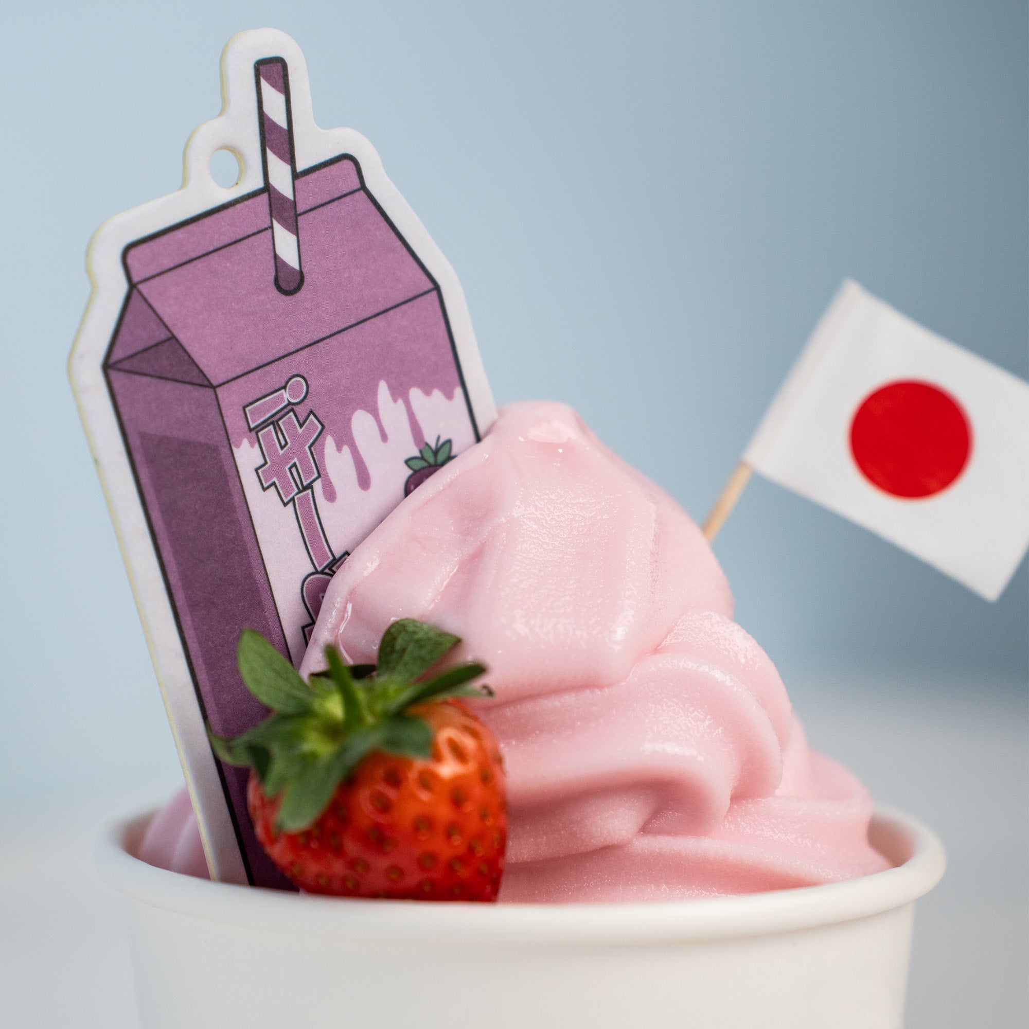 A 'The Milkshake Collection' Air Freshener - Strawberry Milkshake by Blossom Automotive, shaped like pink soft-serve with a strawberry, Japanese flag, and cute purple milk carton stick, set against a light blue background.