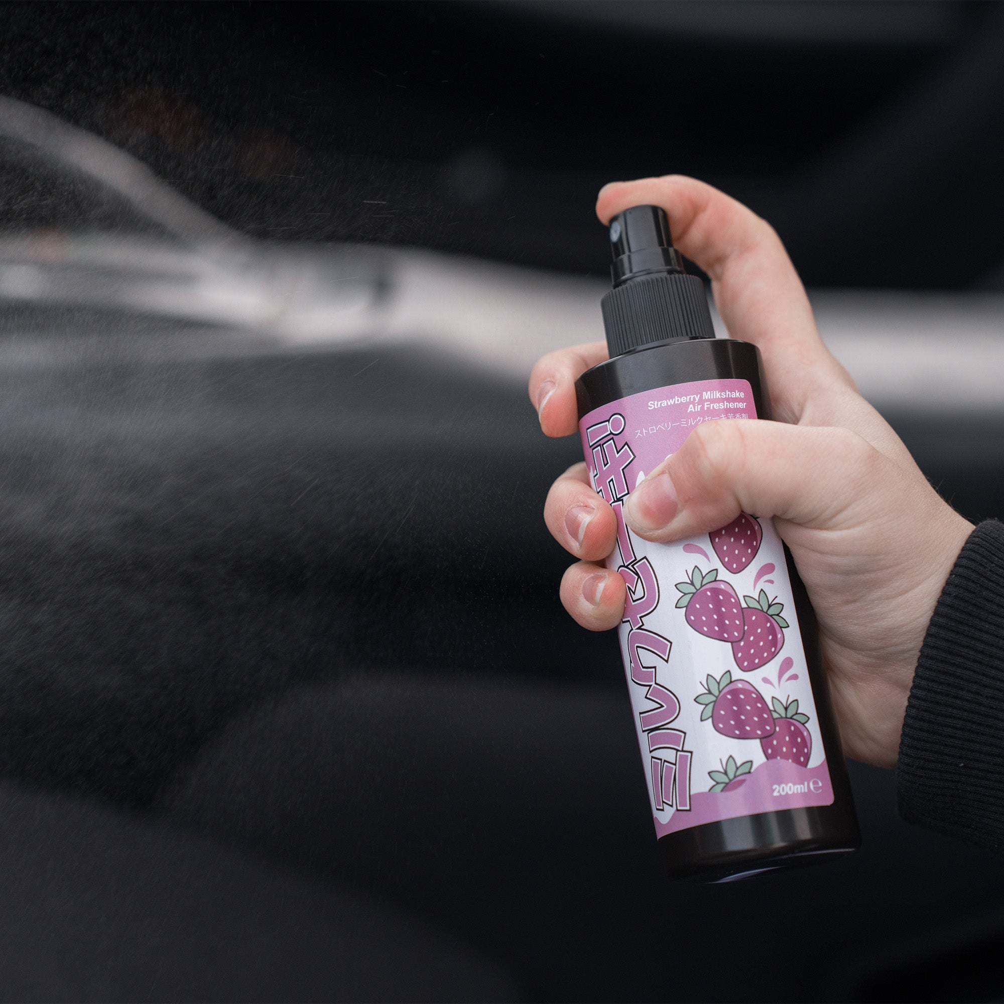 A hand sprays Blossom Automotive's 'The Milkshake Collection' Car Spray Air Freshener (Bundle 2x200ml) in a car. The label features strawberries, and the blurred background suggests use inside a vehicle.