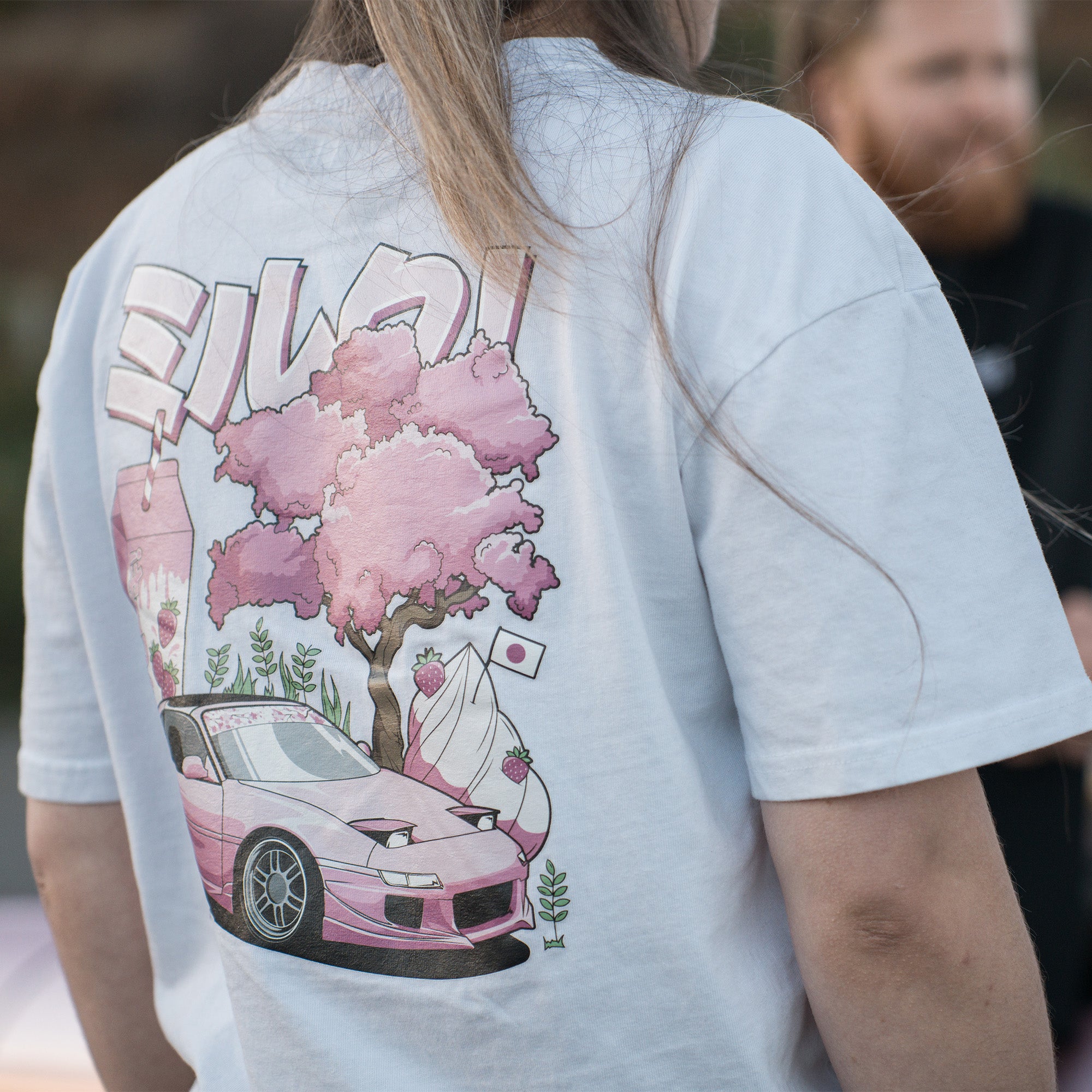 A person outdoors wears The Milkshake Collection Oversized T-Shirt in white by Blossom Automotive, featuring a pink sports car, cherry blossom tree, Japanese elements, and stylized text, with others visible in the background.