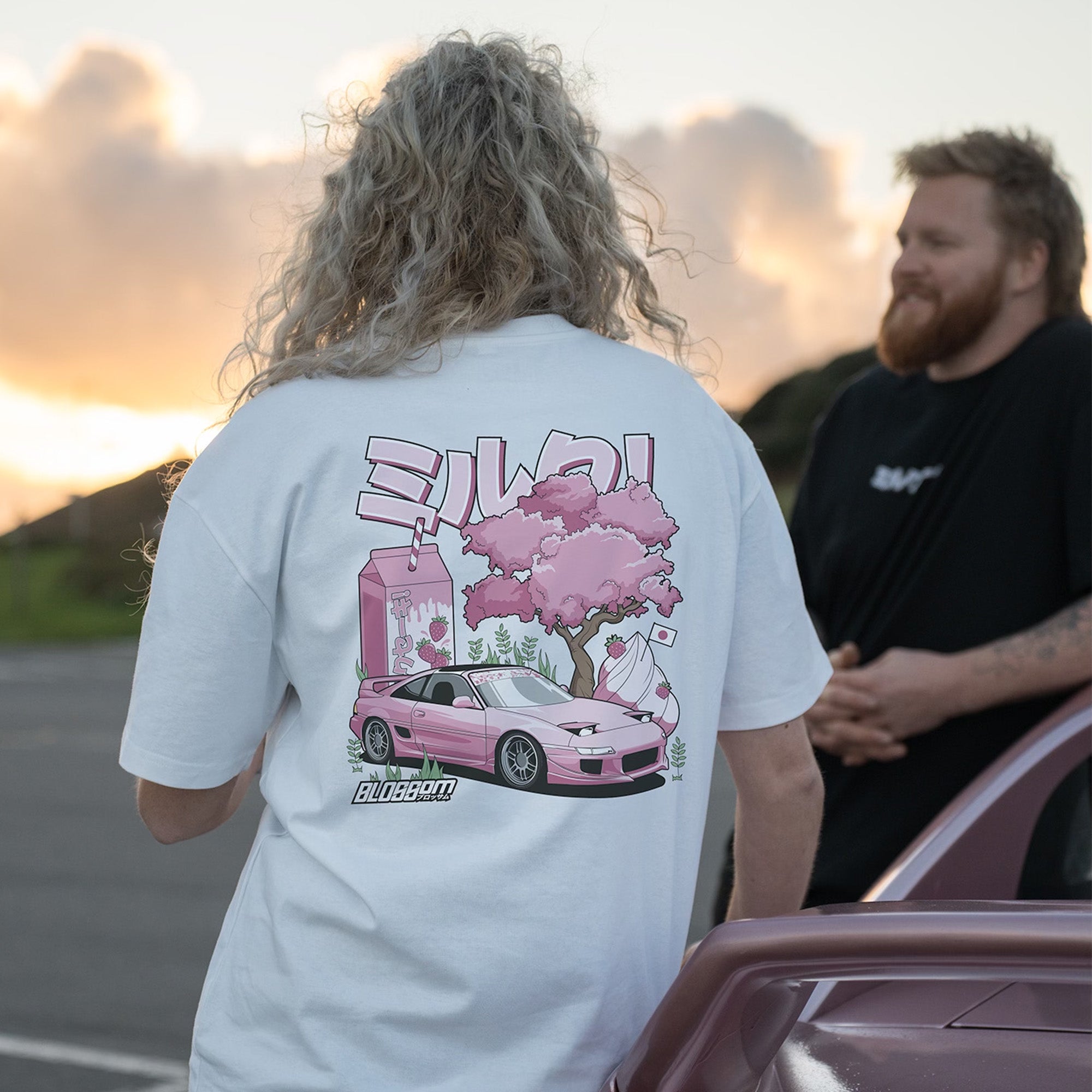 A person with curly blond hair wears Blossom Automotive's The Milkshake Collection Oversized T-Shirt in white, featuring a pink car, cherry blossom tree, and strawberry milk with Japanese text. Another person stands beside them near a parked car at sunset.