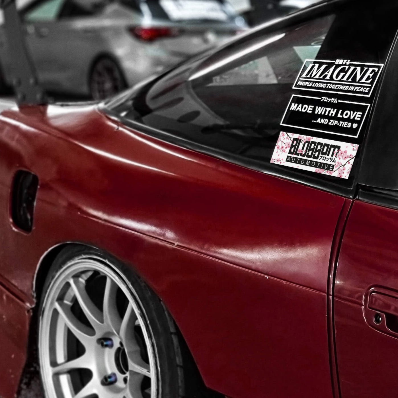 Close-up of a maroon sports car with white wheels. The rear window displays multiple stickers, including the "Made With Love... And Zip-Ties - Die Cut Sticker" by Blossom Automotive. Another car appears blurred in the background.