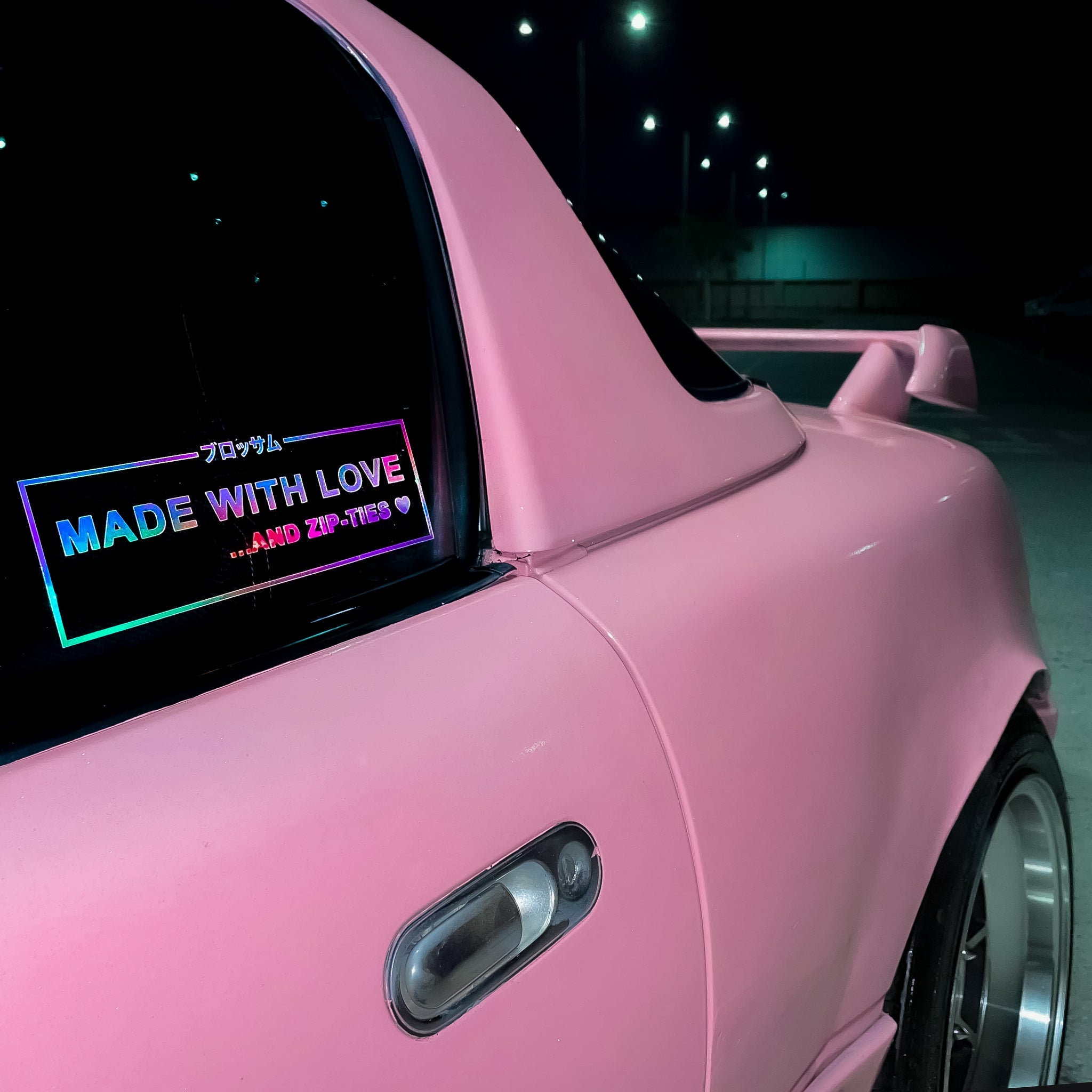 A close-up of a pink car at night shows its rear and a Blossom Automotive "Made With Love... And Zip-Ties - Die Cut Sticker" in holographic colors on the window, with streetlights and a dark sky in the background.