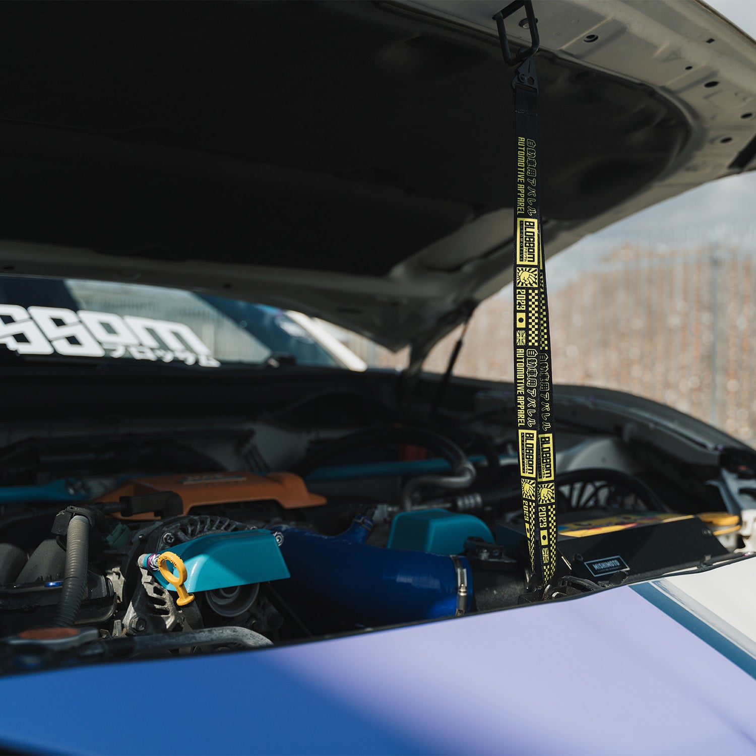 A close-up of a car engine with blue parts features the Blossom Automotive ‘The Mx5 Collection’ Lanyard hanging from the open hood. A white-text windshield decal is partially visible in the background.