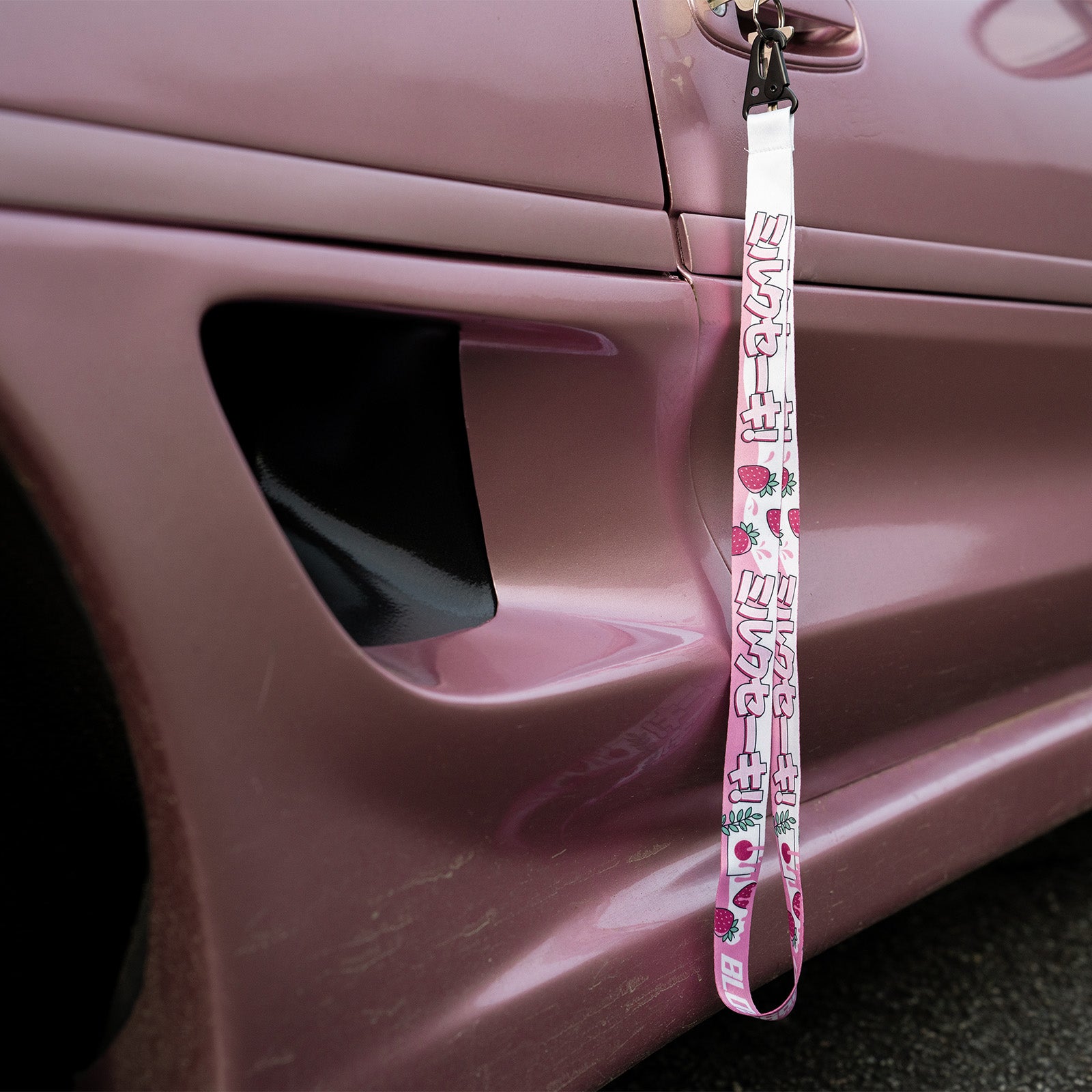 A pink car door with 'The Milkshake Collection' lanyard by Blossom Automotive, featuring pink and green cartoon graphics and “SUPREME,” hanging from the white door handle.