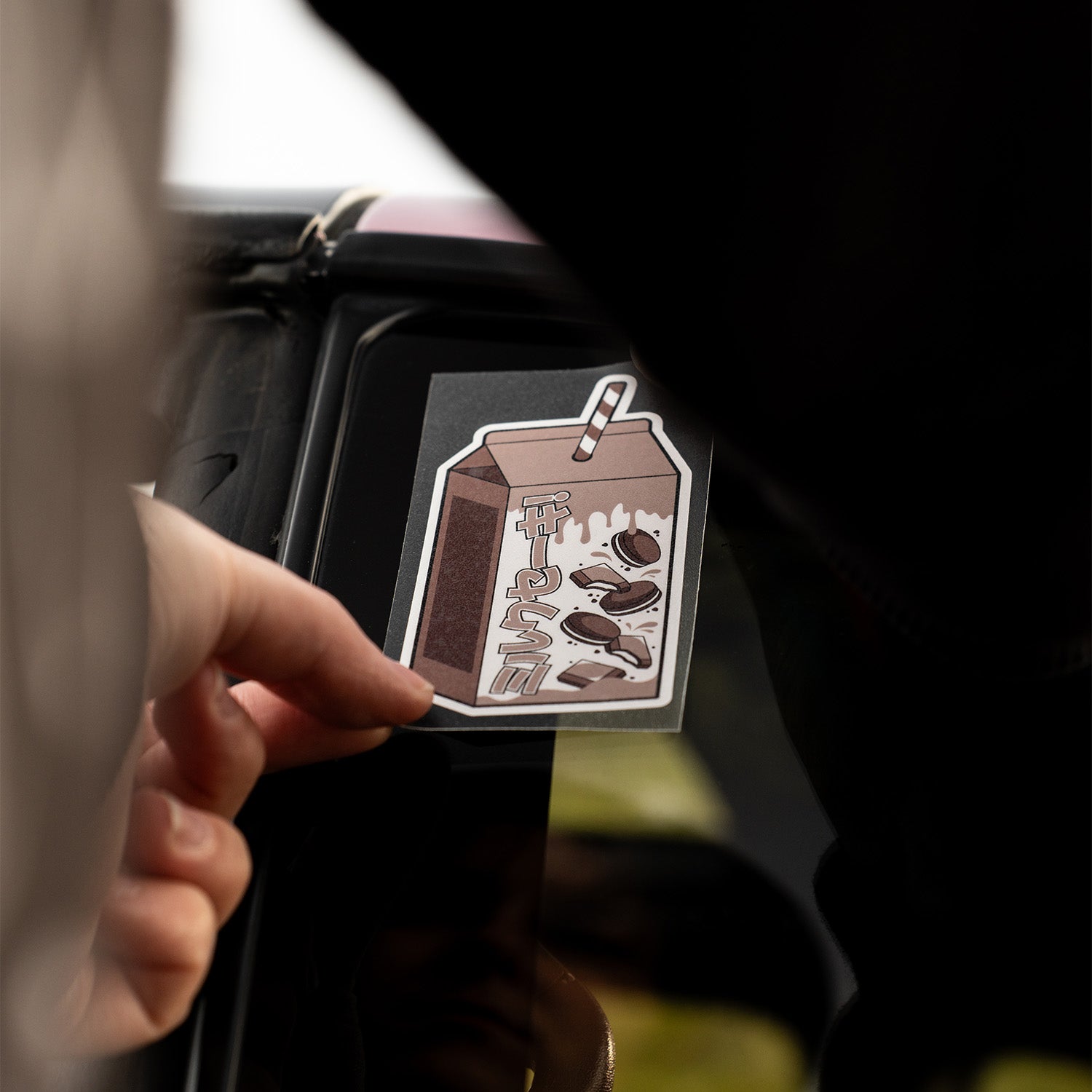A hand places a Blossom Automotive Cookies and Cream Milkshake - Slap Sticker onto a car window.
