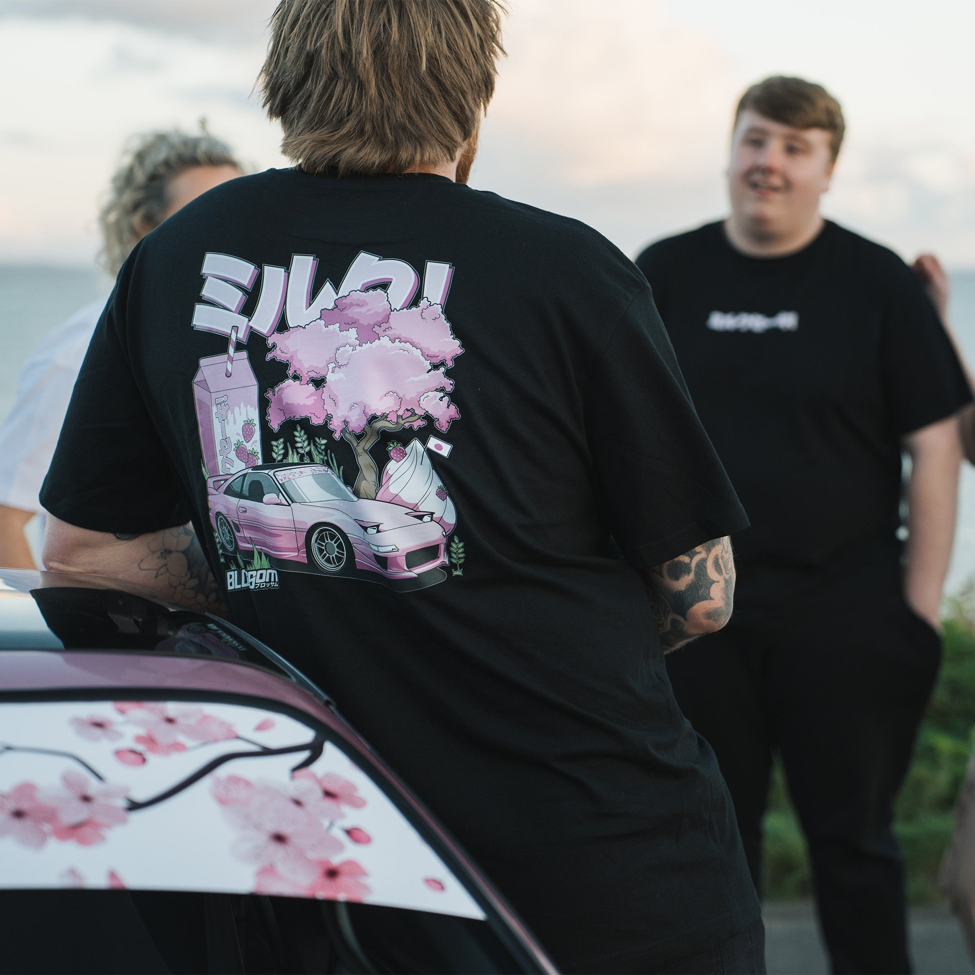 A person wearing a black t-shirt with a graphic design featuring a pink car, strawberry milkshake and cherry blossoms on the back, leaning on a pink car