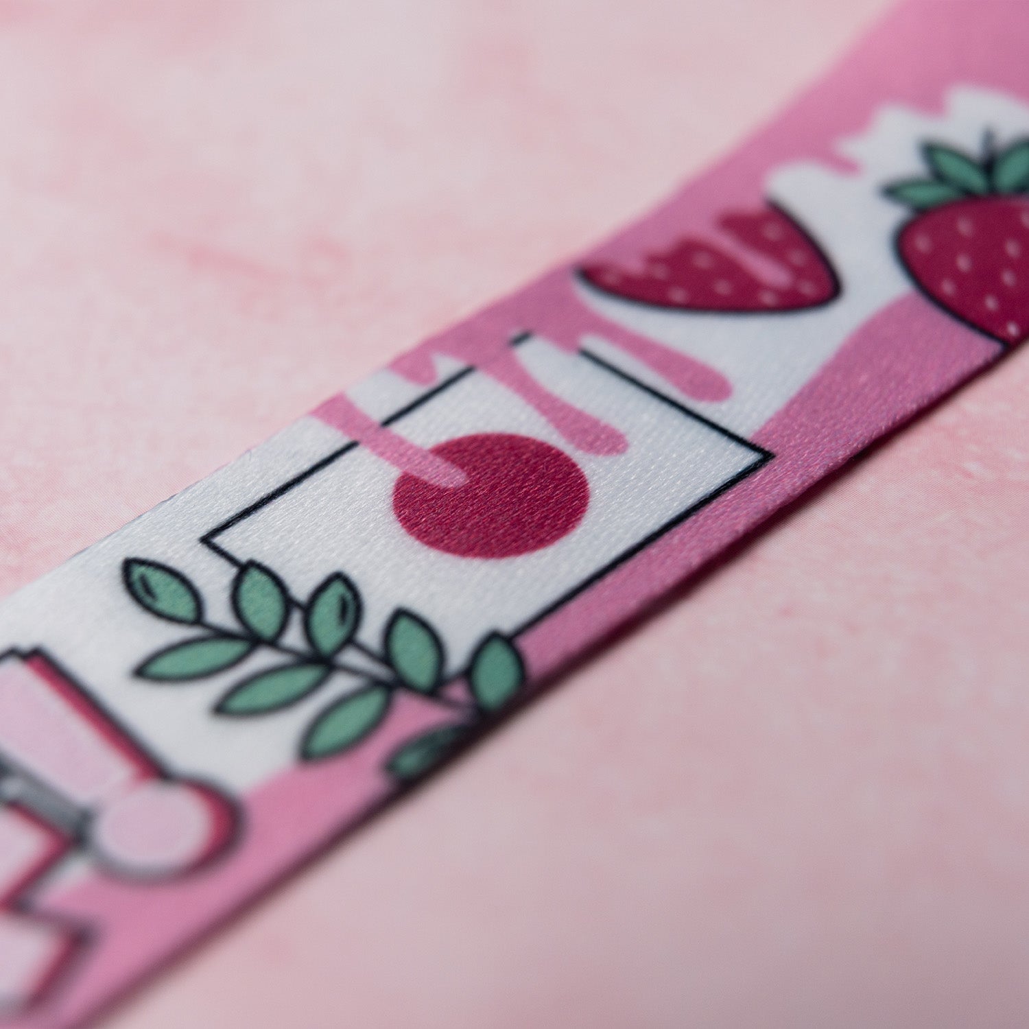 A close-up of Blossom Automotive’s ‘The Milkshake Collection’ Lanyard, showcasing its pink fabric with printed strawberries, leaves, and geometric shapes on a soft pink background.