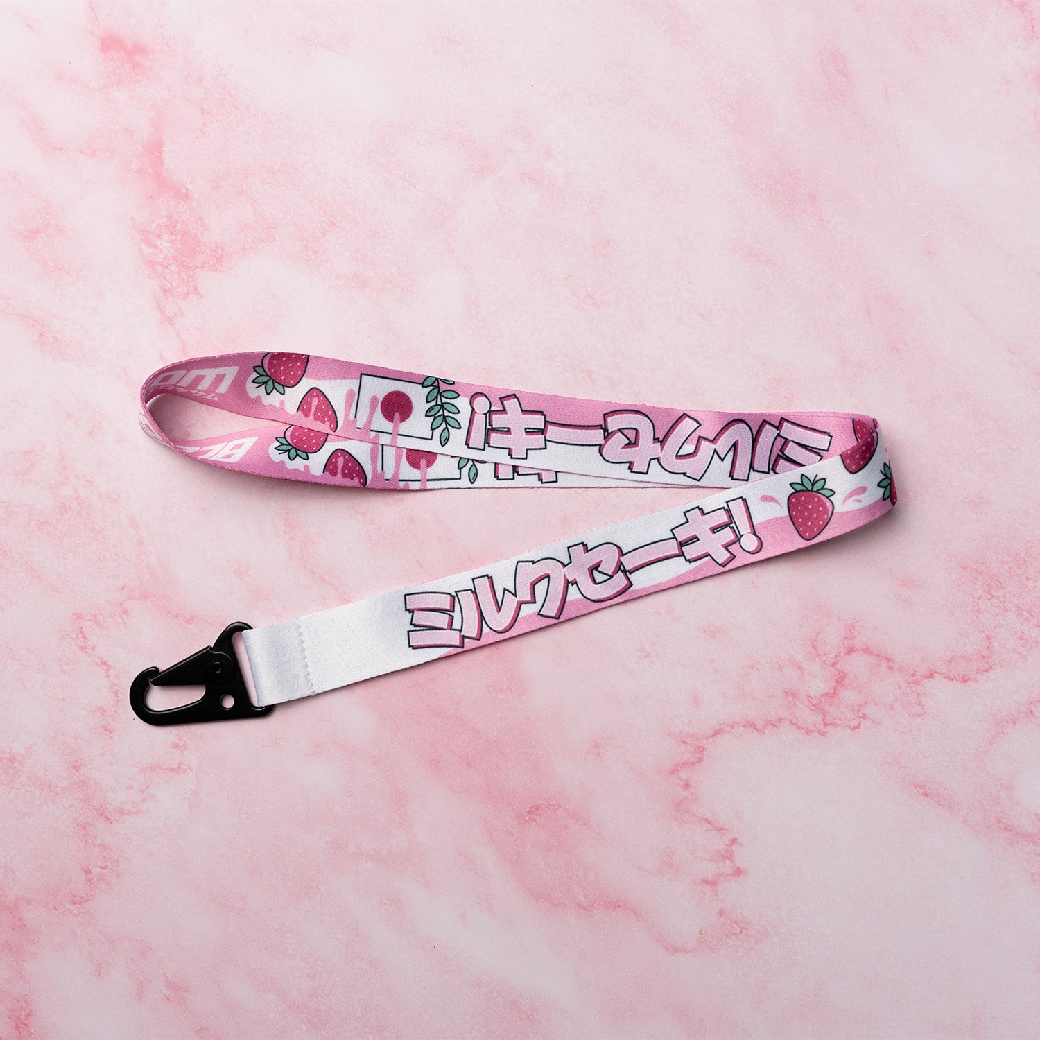 The Blossom Automotive ‘The Milkshake Collection’ Lanyard, featuring pink Japanese characters and cartoon strawberries with a black clip, is displayed on a pink marbled surface.