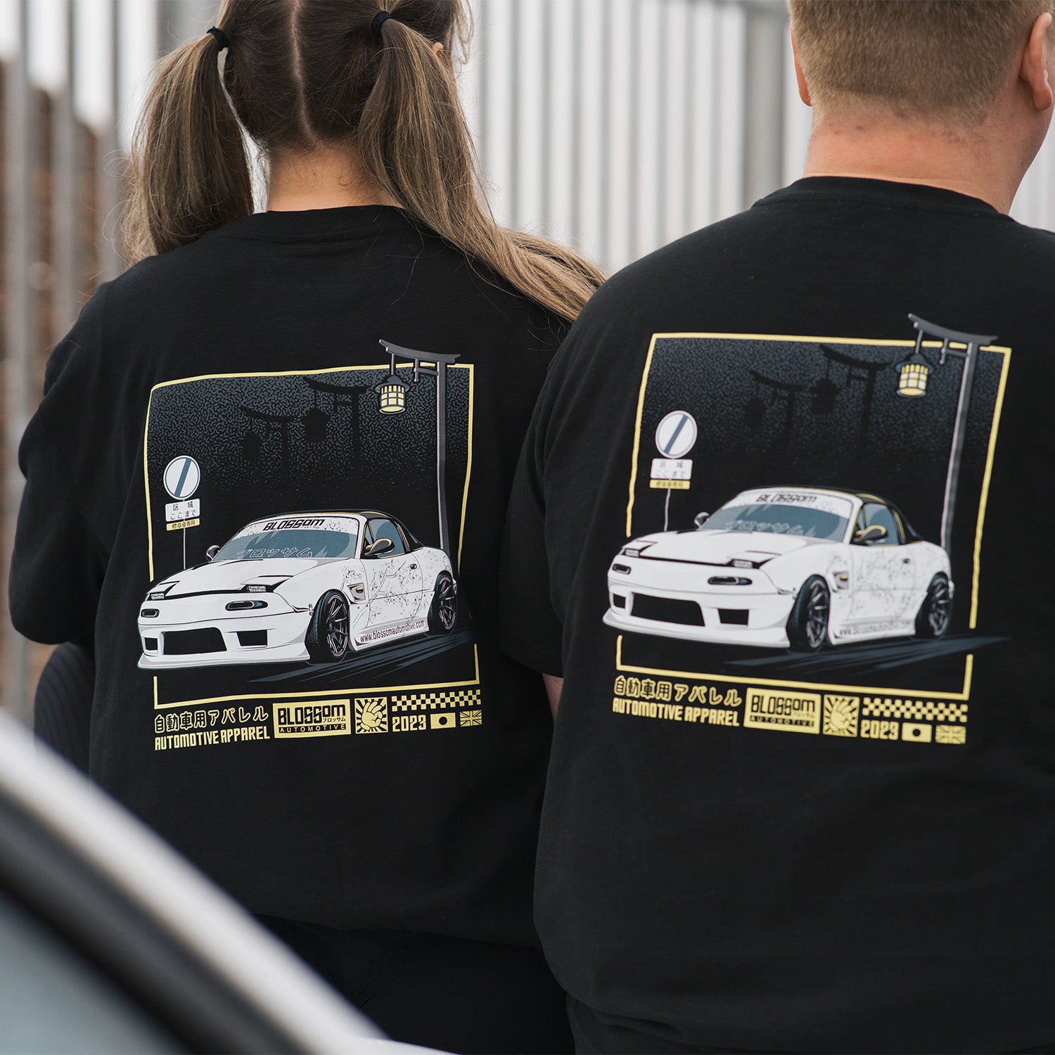 Two individuals sitting on a white mx5, wearing black crewneck t-shirts with a yellow graphic design on the back, featuring a white mx5.