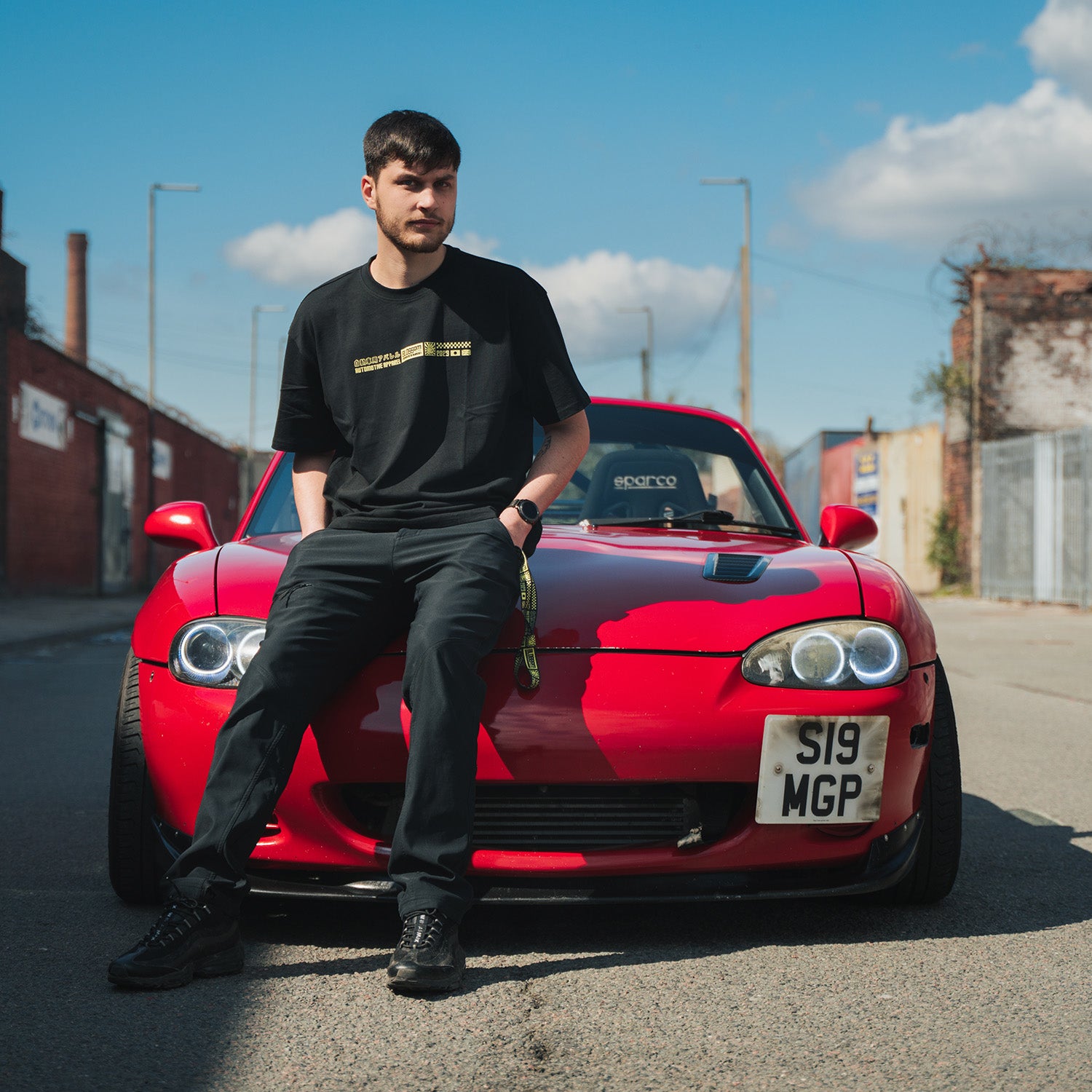A person sitting on a red mx5, wearing a black crewneck t-shirt from the mx5 collection