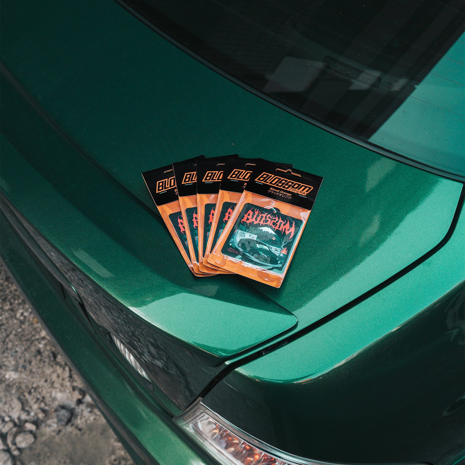 Five 'The Skate Collection' Air Fresheners in Blood Orange by Blossom Automotive, each featuring a car graphic, are displayed on the trunk of a shiny green car outdoors in daylight.