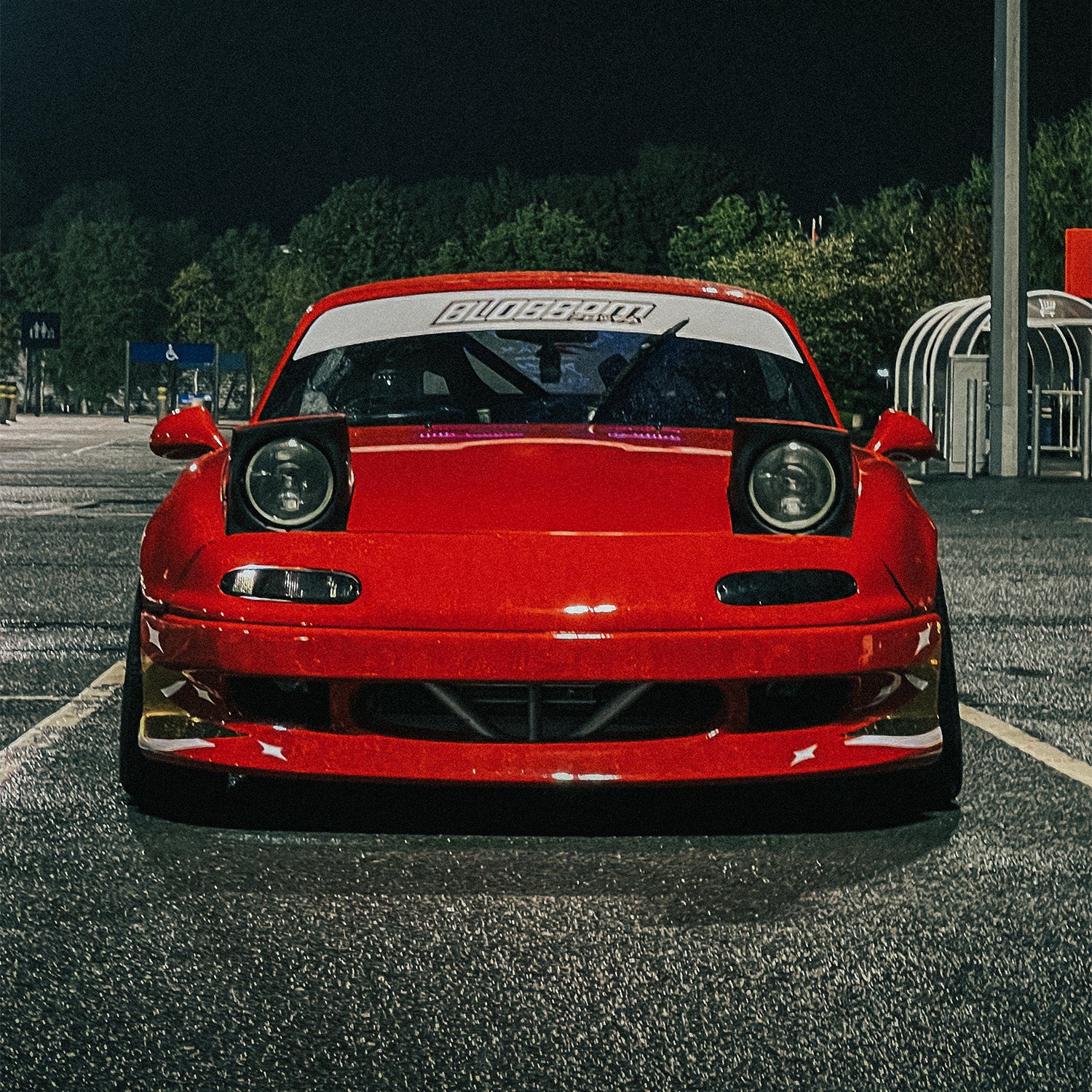 A red Mazda MX-5 Miata with pop-up headlights up, parked in a lot at night, features an aftermarket front lip and a Blossom Automotive "Blossom - Sunstrip Banner" on the windshield. Trees and parking structures are visible in the background.