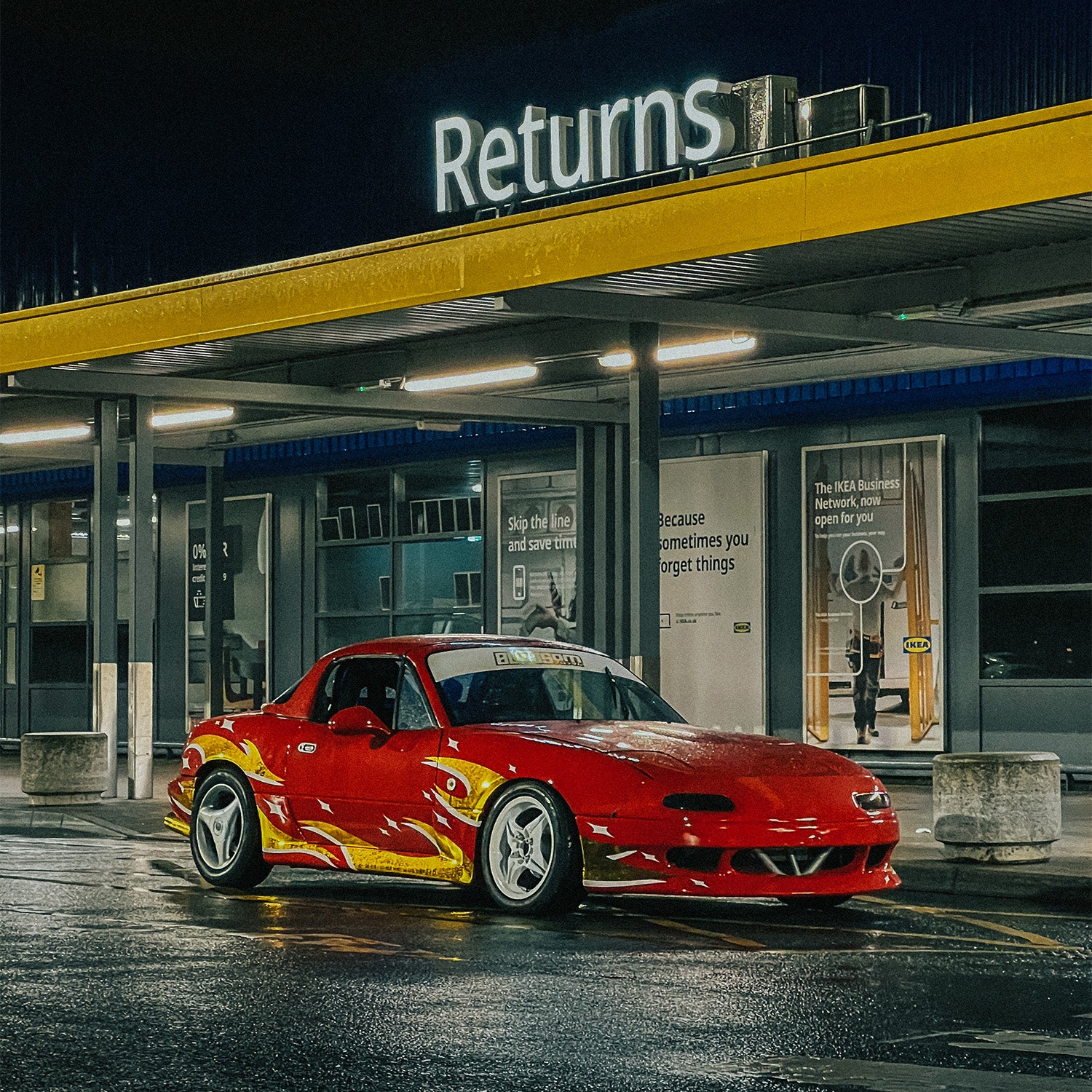 A car with a Blossom - Sunstrip Banner by Blossom Automotive is parked at night under a yellow-lit “Returns” sign outside a glass-fronted building. Wet ground reflects the lights and the banner.