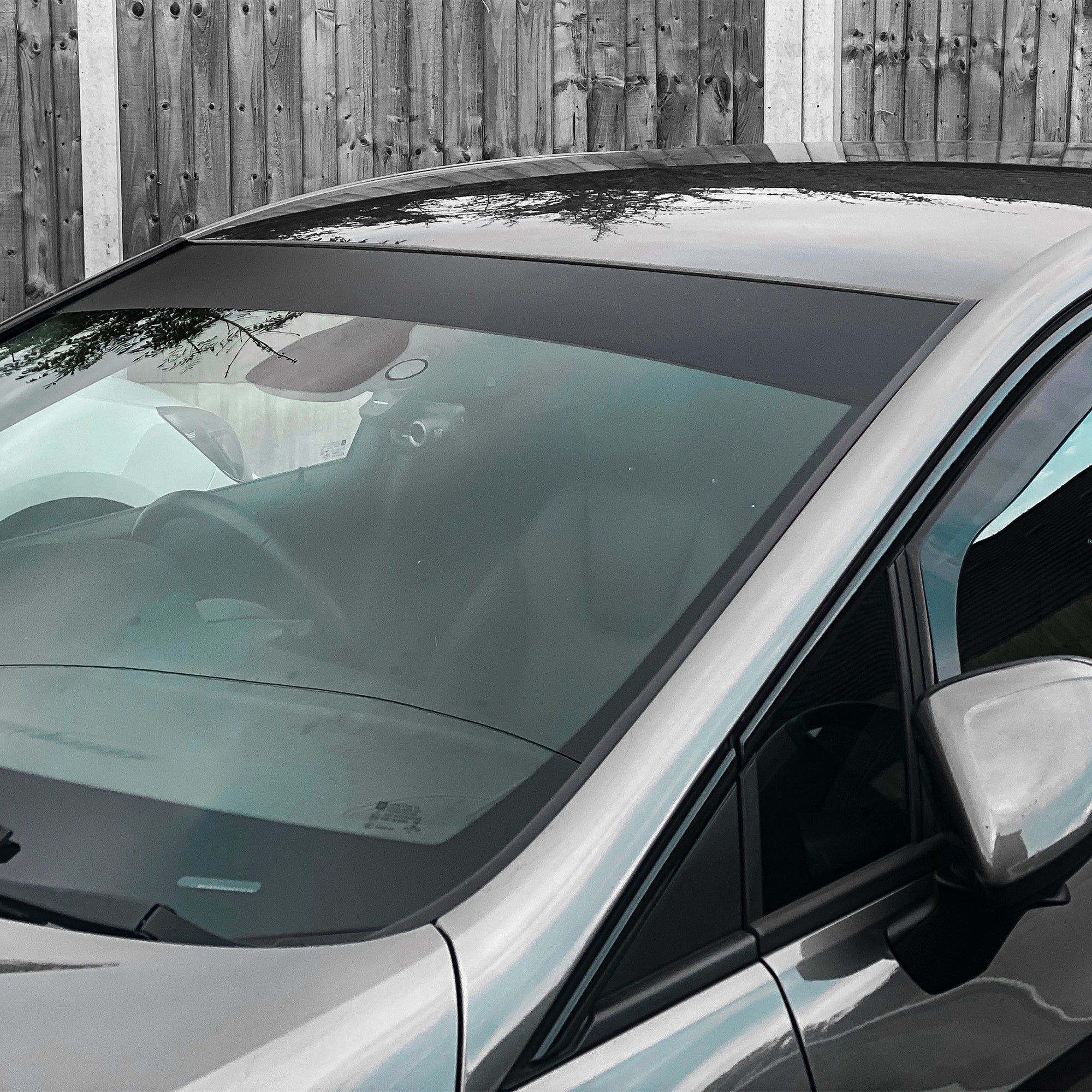 A close-up of a silver car with a Blossom Automotive Plain Black Sunstrip (Gloss/Matt) on the windshield, parked by a wooden fence. The car's interior and steering wheel are faintly visible through the glass.