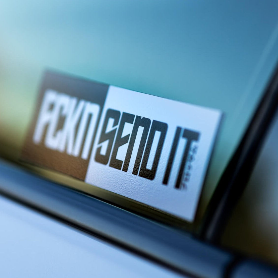 A Blossom Automotive FCKN Send It - Slap Sticker in black and white is angled on a car window, showing off its bold text with a blurred background seen through the glass.