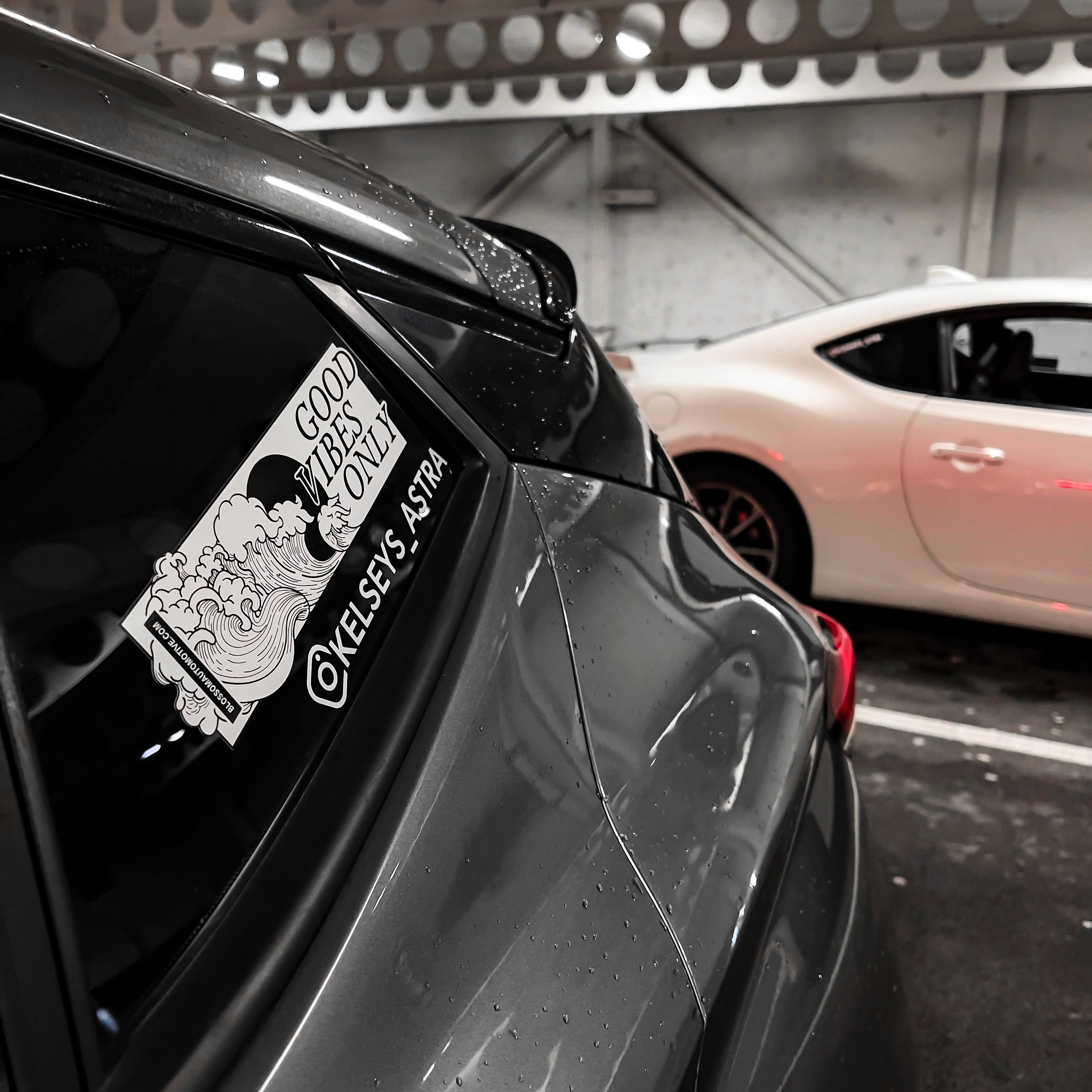 Close-up of a car window showing the Blossom Automotive “Good Vibes Only - Slap Sticker” with waves, clouds, and “Kelsea S. Astra.” A white car is parked beside it in a covered parking area.