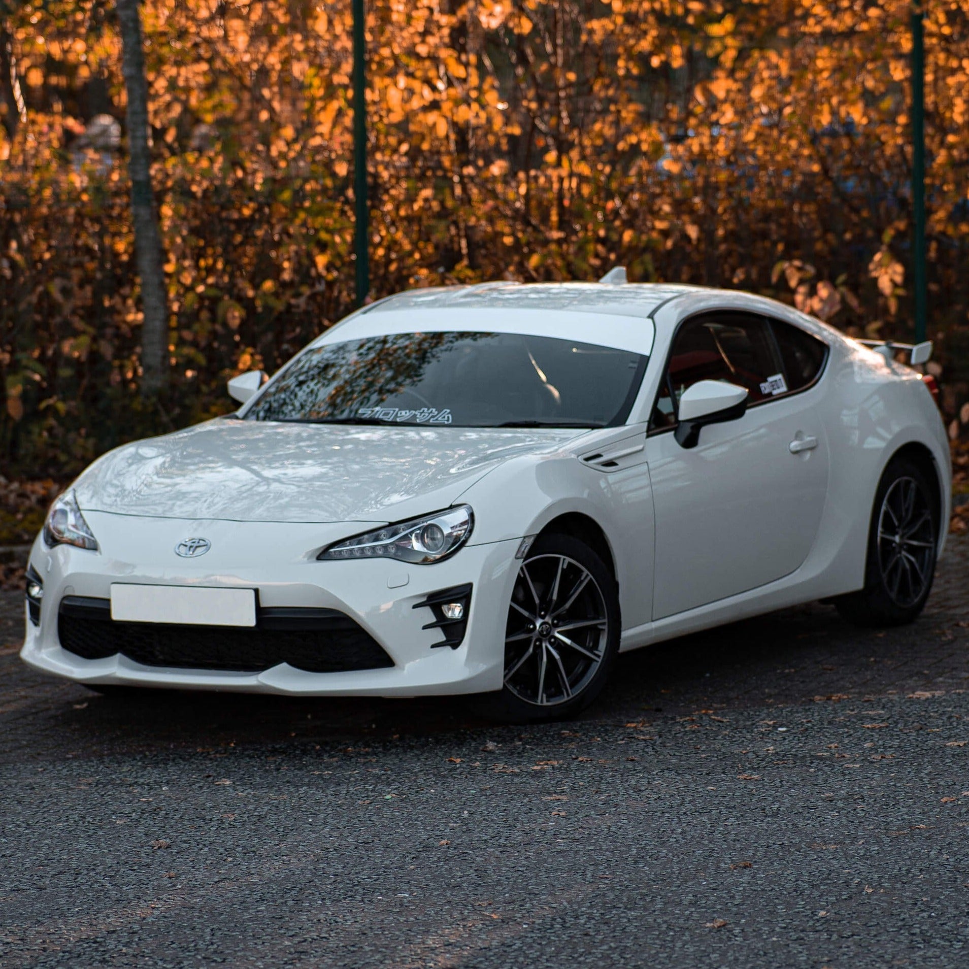 A white sports car with a Blossom Automotive Plain White Sunstrip (Gloss/Matt) is parked on a street lined with autumn-colored trees and fallen leaves. The car features sleek lines, black wheels, and tinted windows.