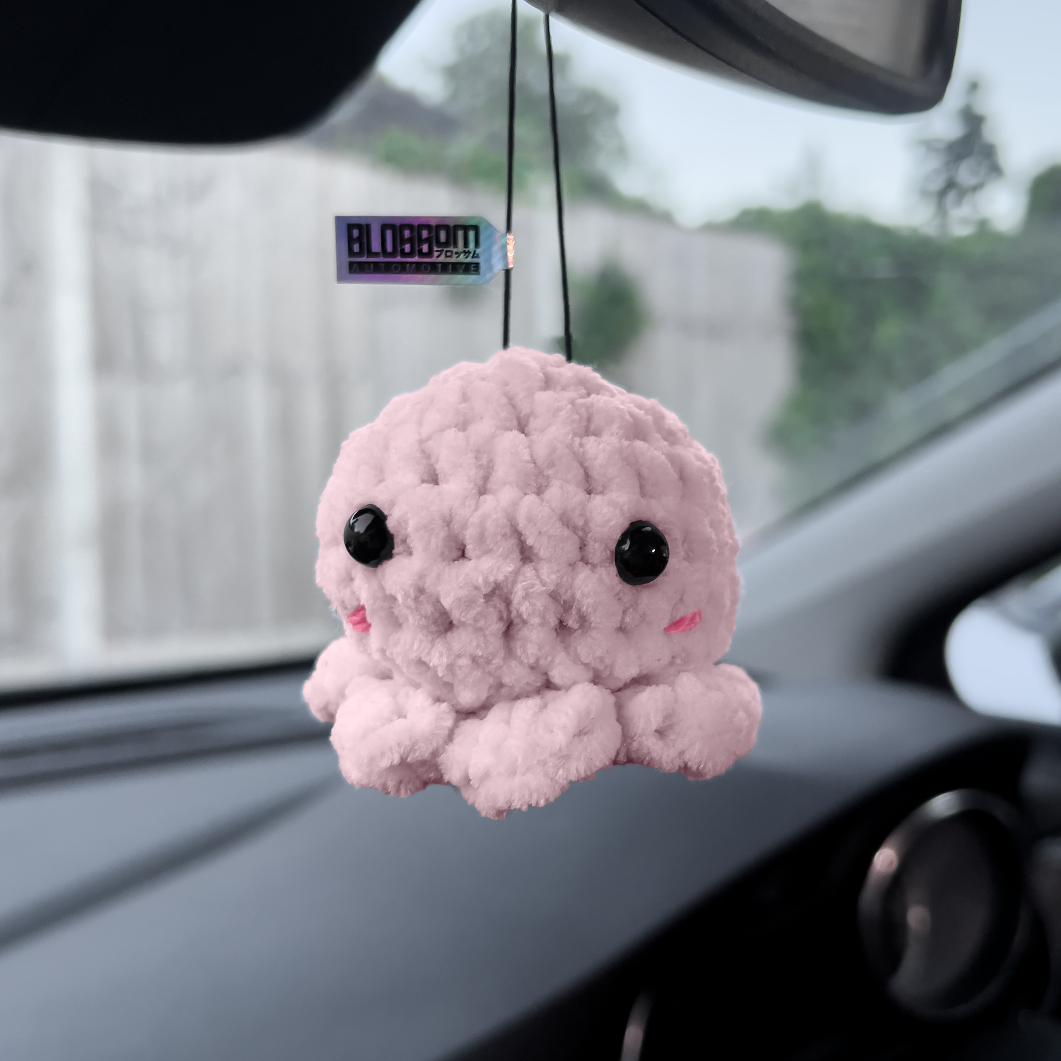 The Squishy Pink Octopus - Car Mirror Hanger by Blossom Automotive is a small, plush pink octopus with black eyes and a tiny smile, hanging from your rearview mirror against a blurred background of fence and greenery.