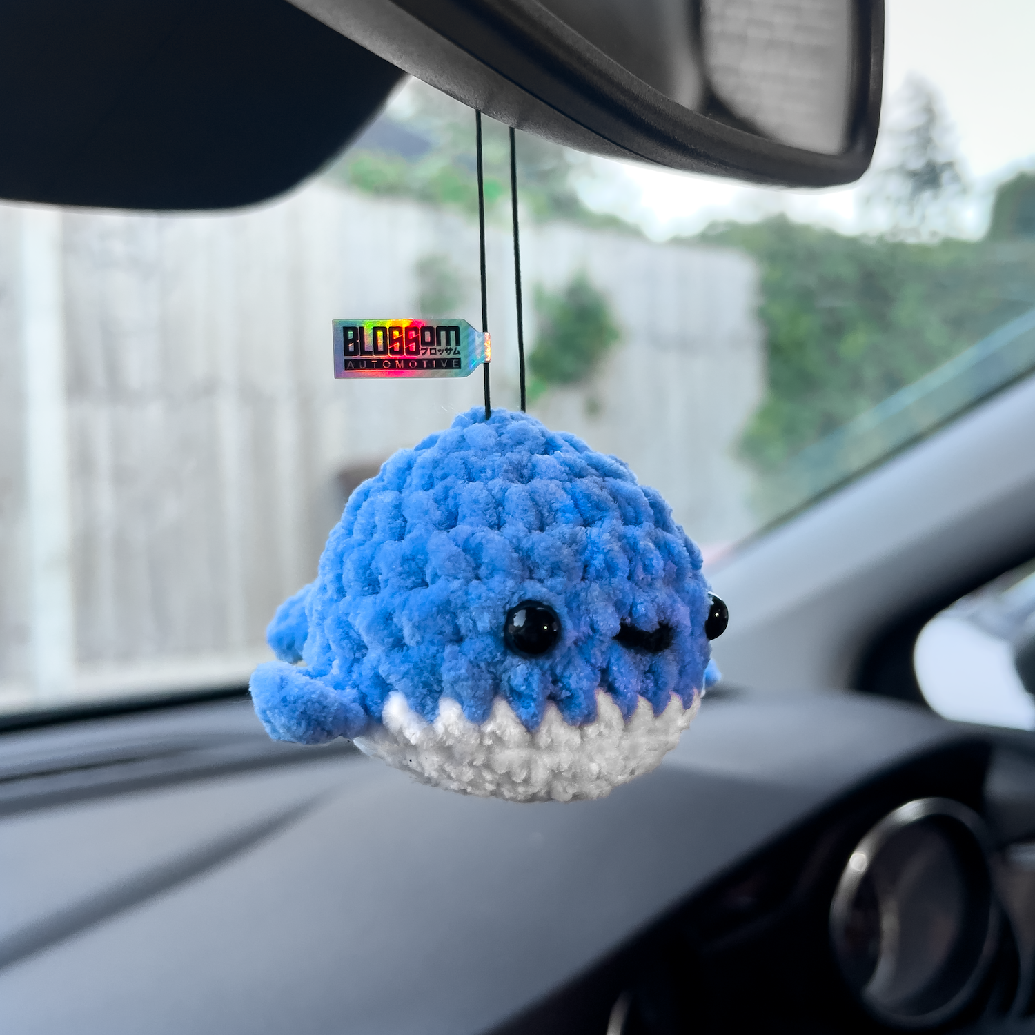 The Squishy Blue Whale - Car Mirror Hanger by Blossom Automotive, a soft blue plush with black eyes and a white belly, hangs inside the car. A colorful tag is attached, and greenery and a wooden fence are visible outside the window.
