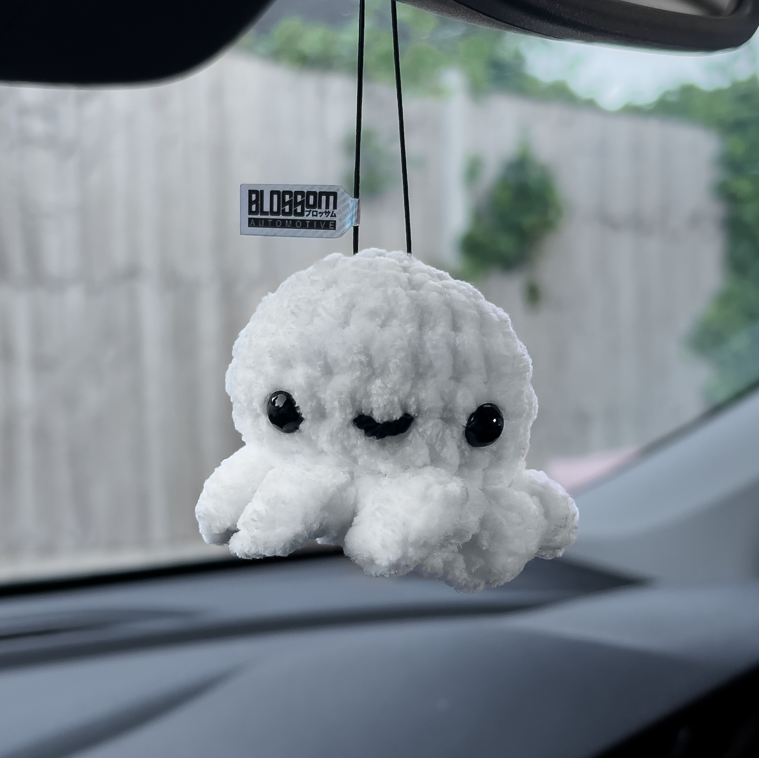 The Blossom Automotive Squishy White Octopus - Car Mirror Hanger is a cute plush toy with black eyes and a smiling face, hanging by a string inside the car. A blurred fence and green foliage are visible outside the window.