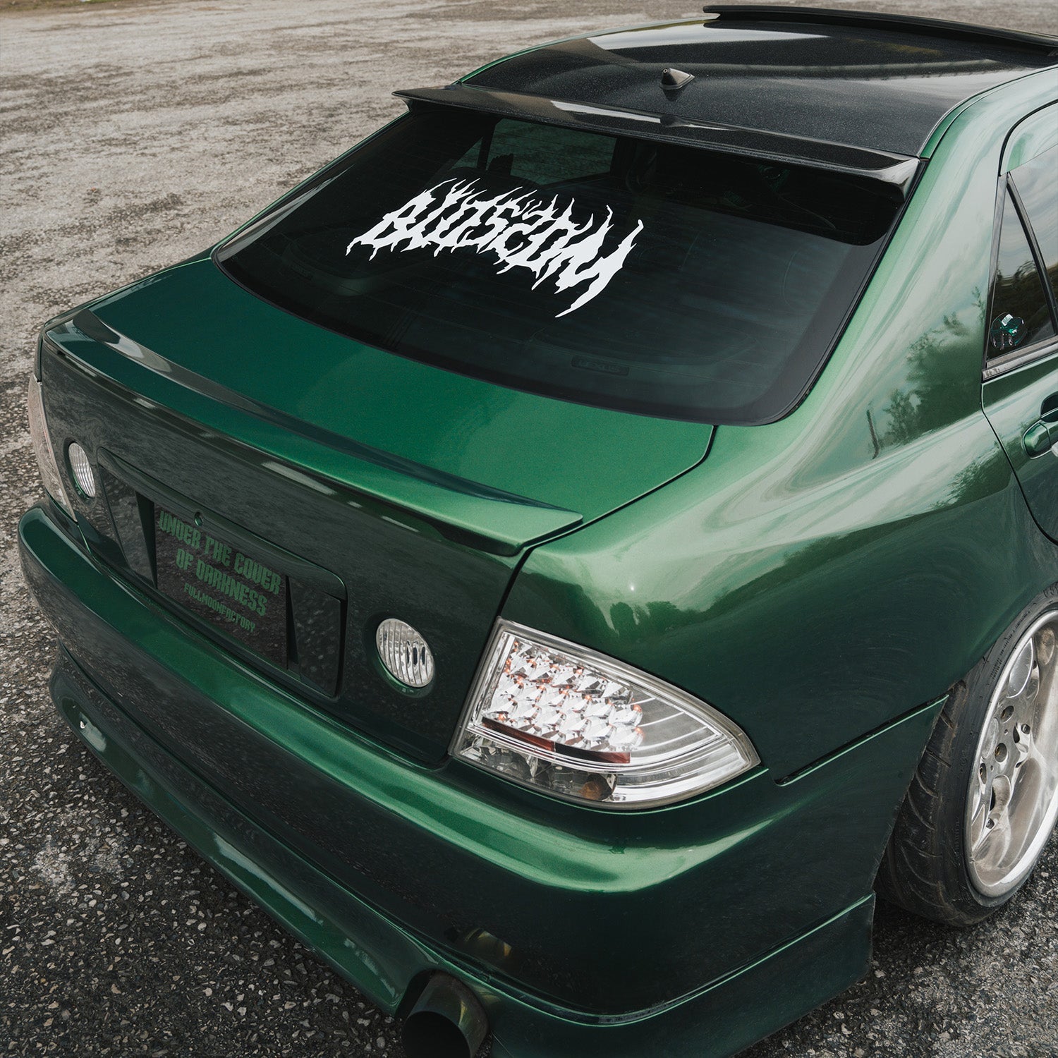 A white decal with jagged, flame-like edges spells BIOSROM on the rear windshield of a green car, reflecting clouds and sky.